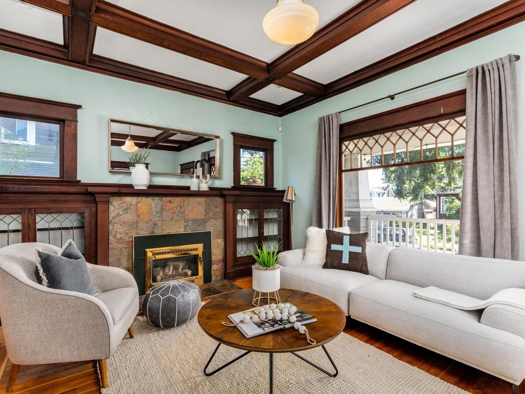 A cozy living room features a white sofa, a gray armchair, and a round wooden coffee table. Theres a fireplace with a stone surround, built-in shelving, and a large window with gray curtains. The room has wooden ceiling beams and a light fixture.