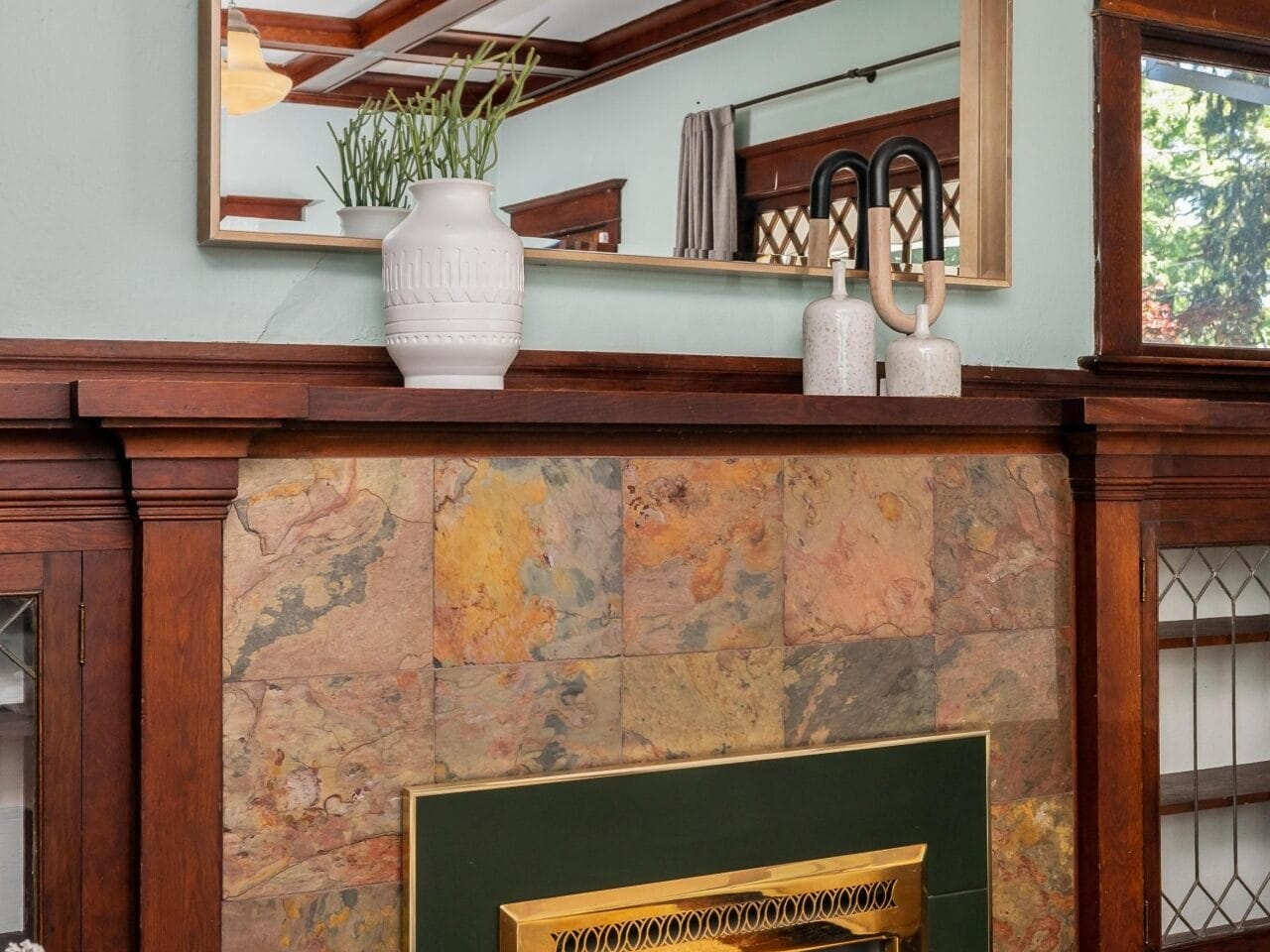 A cozy living room with a decorative fireplace framed by a dark wood mantel. Above the mantel is a large mirror. A white vase with green plants and two sculptural pieces adorn the mantel. The wall is painted light blue-green.