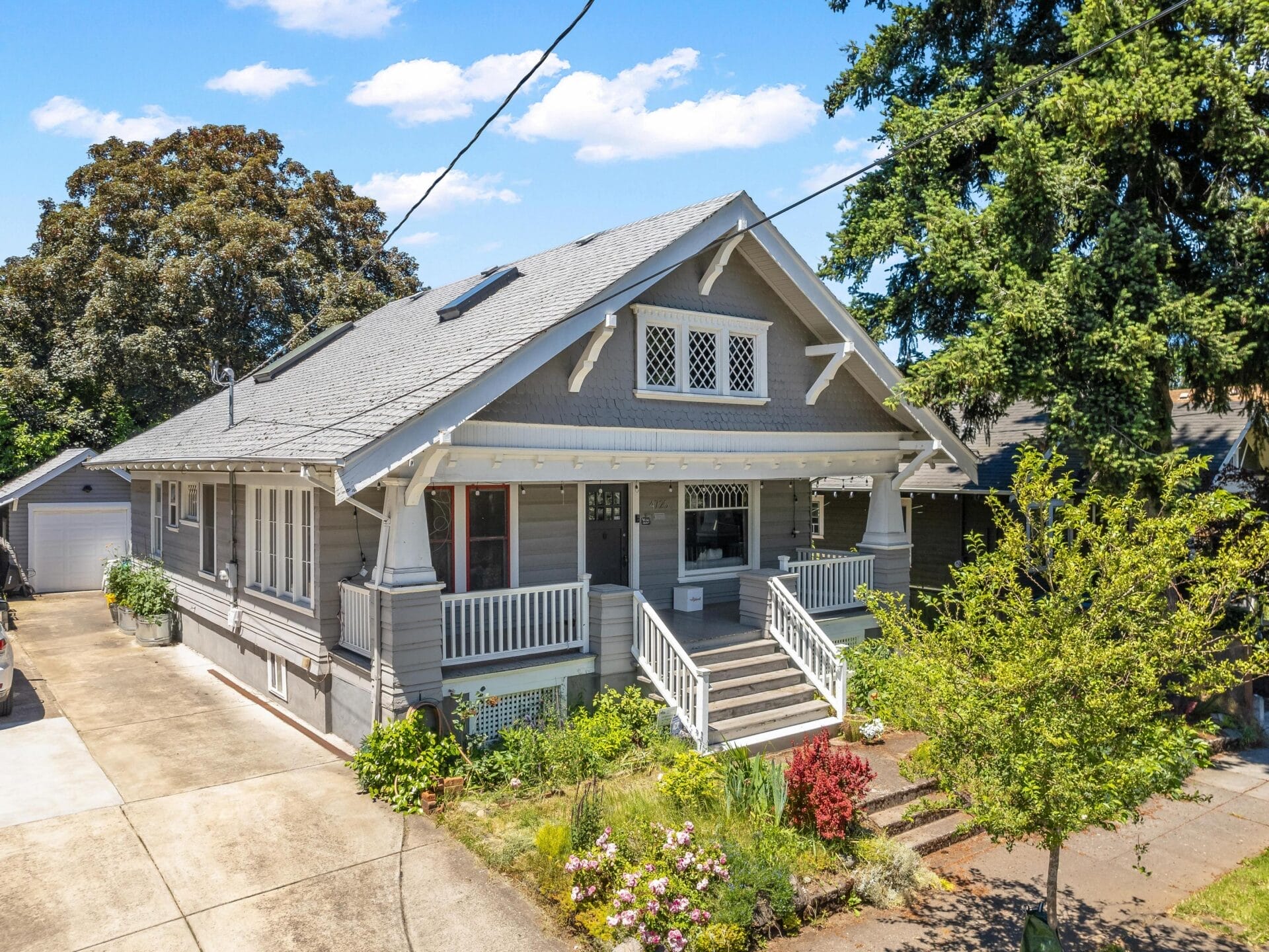 A charming gray house with white trim features a spacious front porch, gabled roof, and lattice windows. It is surrounded by trees and a garden with colorful flowers. A driveway runs alongside the house, leading to a garage. Bright blue sky above.