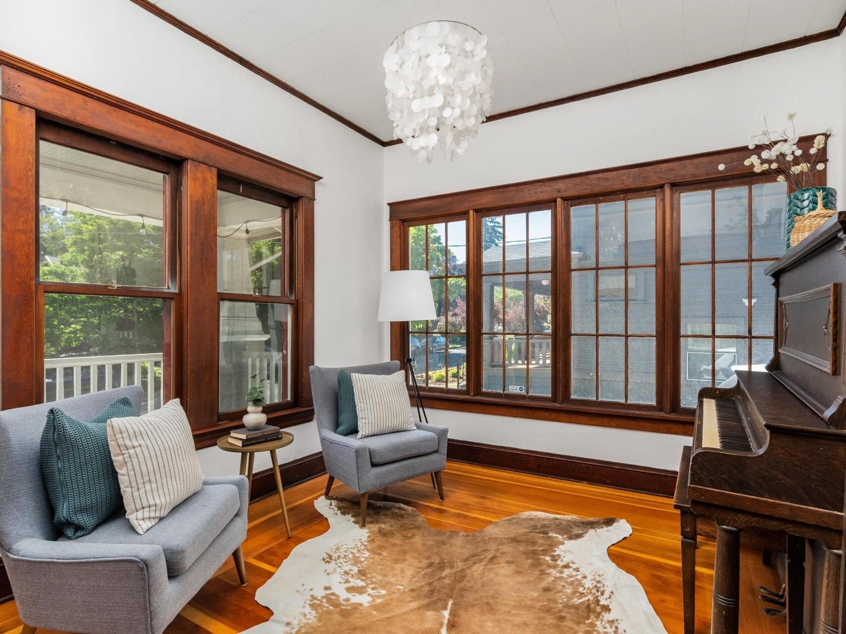 A cozy room with large wooden-framed windows, two gray armchairs with cushions, a small side table, and a piano. A cowhide rug lies on the polished hardwood floor under a modern chandelier.