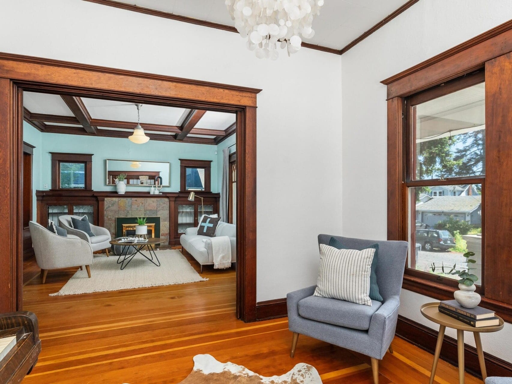 A cozy living room featuring wooden floors and dark wood trim. It includes a blue armchair, a small side table, and a view into a larger room with sofas, a fireplace, and a chandelier. Bright natural light streams through large windows.