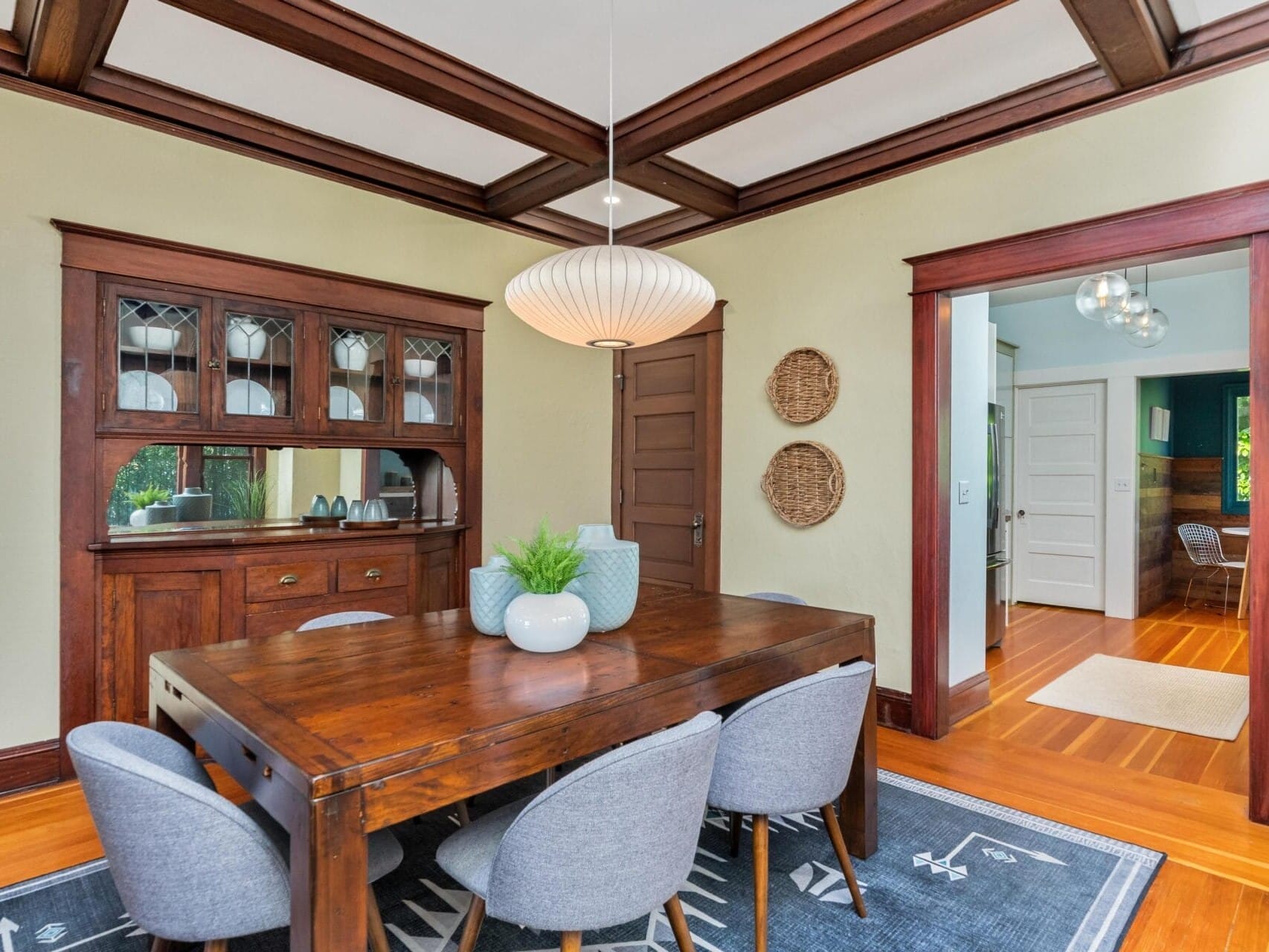 A dining room with a wooden table, six gray chairs, and a modern pendant light. The room features wooden trim, a built-in cabinet with glass doors, and decorative items like vases and wall hangings. Warm lighting enhances the wooden flooring.