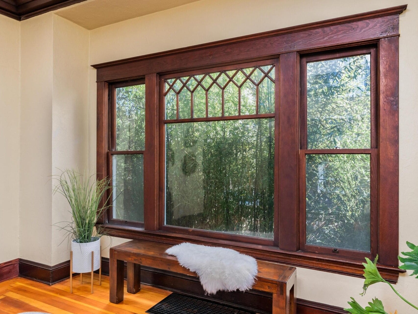 A cozy room featuring a large decorative window with wooden trim. In front of the window, theres a wooden bench adorned with a white sheepskin rug. A potted plant is placed nearby, adding a touch of greenery to the space.