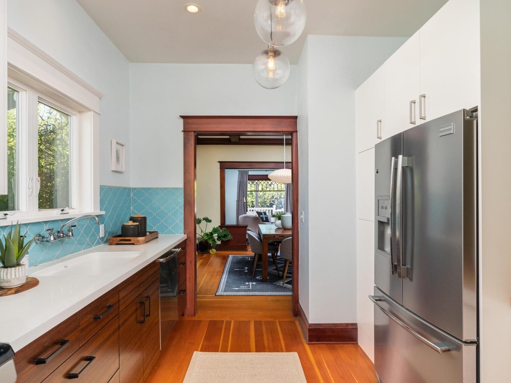 A modern kitchen featuring wooden cabinets, a stainless steel refrigerator, and teal backsplash tiles. The space is well-lit with pendant lights, a large window, and opens to a dining room with a wooden floor and table visible in the background.