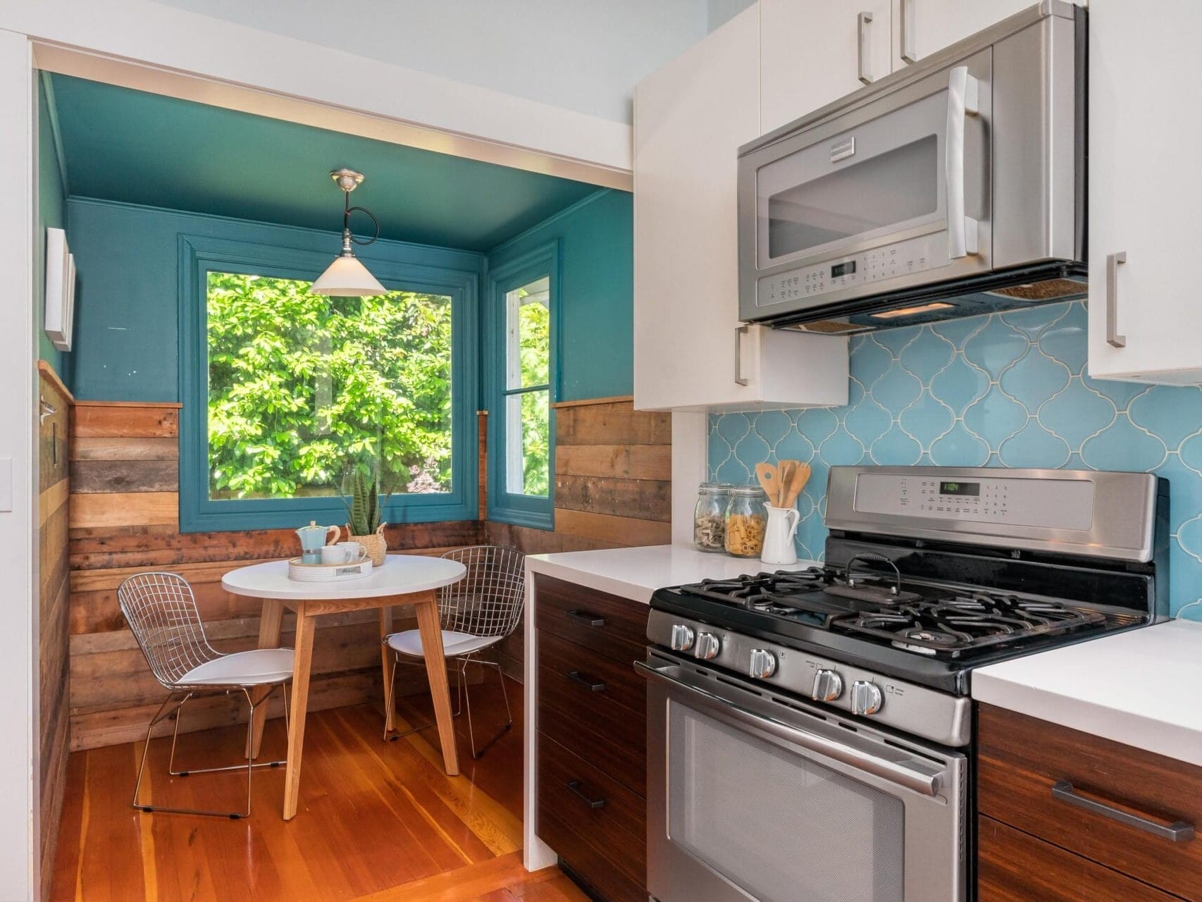 A modern kitchen featuring a gas stove, stainless steel microwave, and white countertops. In the adjacent dining nook, theres a small white table with two wire chairs by a window, and turquoise walls with wooden accents.