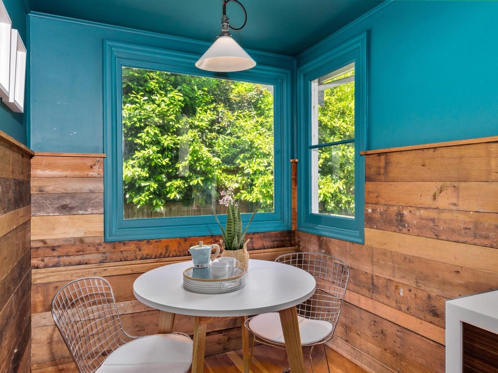 A cozy nook with a round white table set for tea, including a teapot, cups, and saucers. It is surrounded by mismatched wooden walls and bright blue accents. Large windows offer a view of lush greenery outside, and modern wire chairs complete the scene.