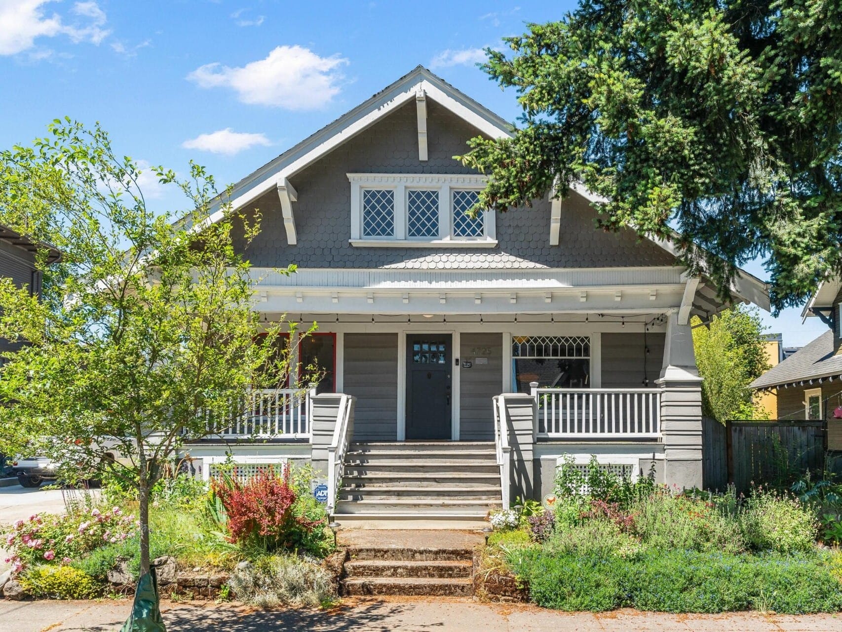 Charming two-story gray craftsman house with a gabled roof and a front porch. It features decorative windows and lush landscaping. A tree to the right partially shades the scene, under a clear blue sky.