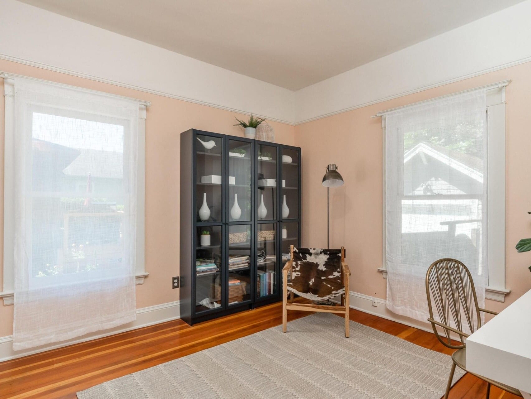 A cozy, light-filled room with wooden floors and a minimalist design. It features a glass-front cabinet with decor, a modern lamp, a patterned chair, a large plant, and a white desk. Two sheer-curtained windows let in natural light.