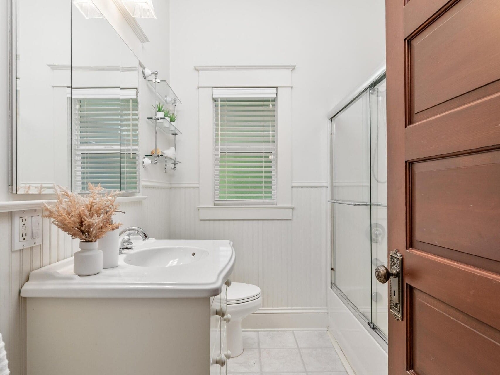 A small, brightly lit bathroom with a white sink, a toilet, and a shower with sliding glass doors. A wooden door is partially open, and two windows with blinds are above the toilet. A small plant and dried flowers decorate the space.