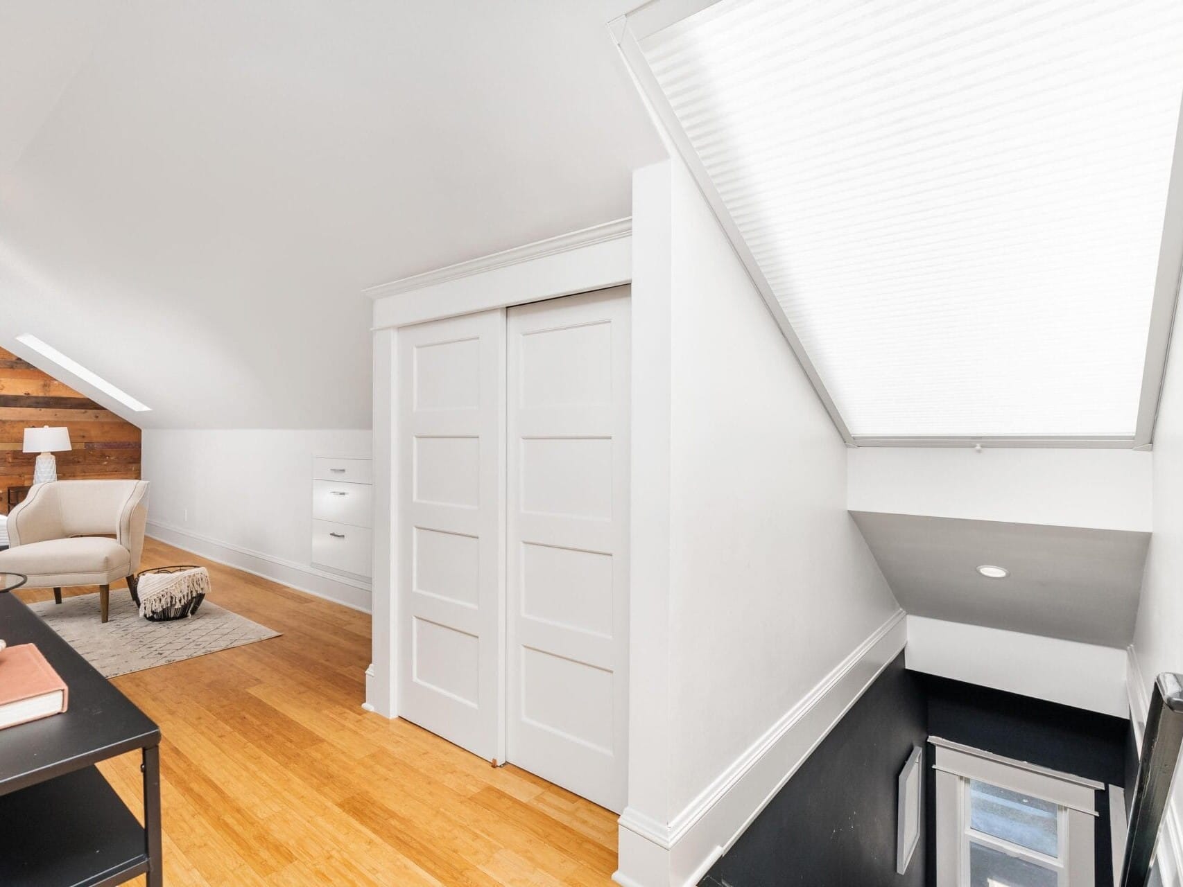 A bright attic space with white walls and a sloped ceiling. It features a seating area with a cushioned chair, hardwood floors, and a small rug. There are built-in closets with sliding doors and a skylight illuminating the staircase.