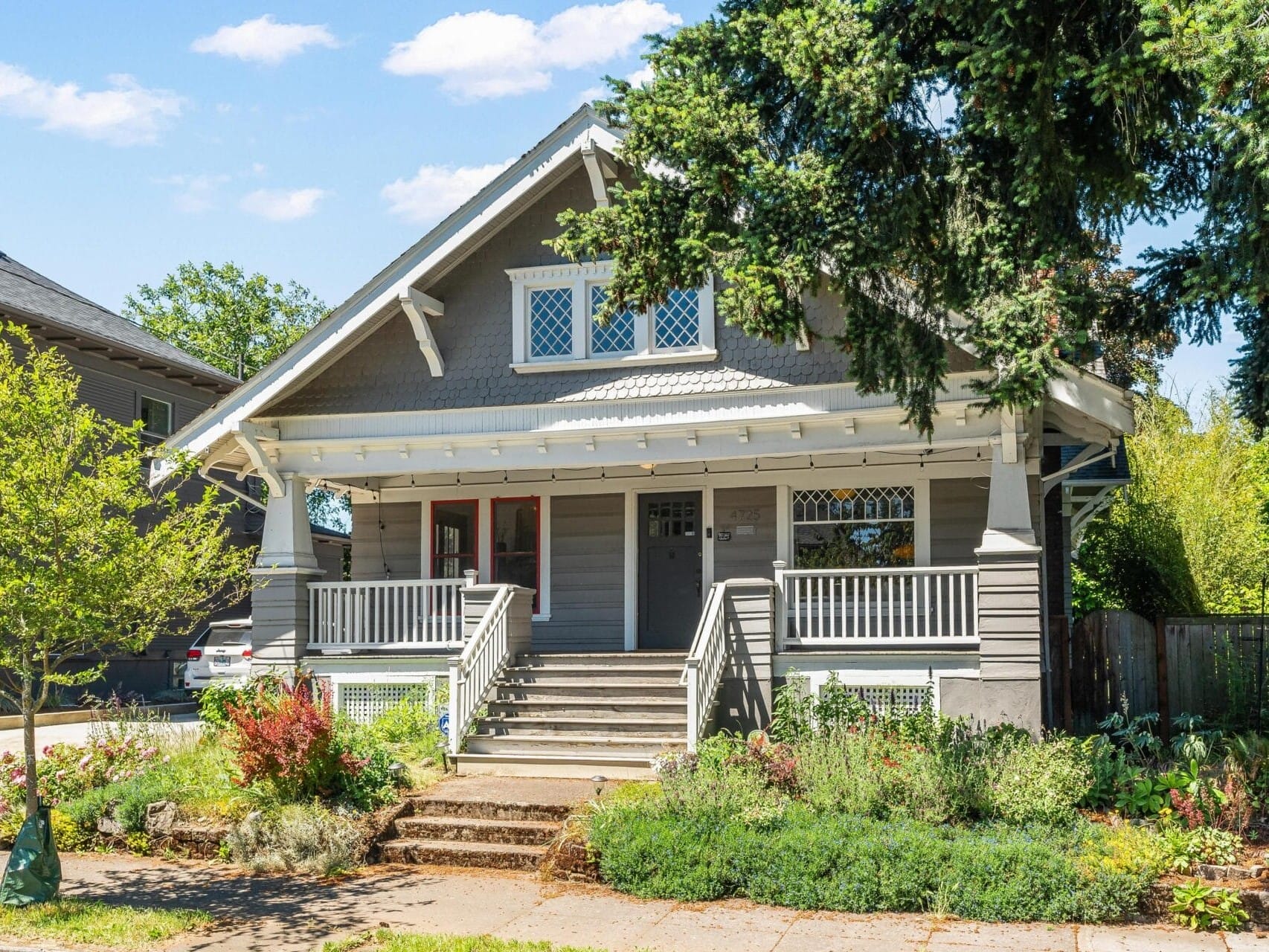 A gray suburban house with a front porch and steps leading up to it. The porch is adorned with white railings and surrounded by lush green plants and shrubs. The house has a gabled roof and windows with lattice designs. A tree is visible on the right.