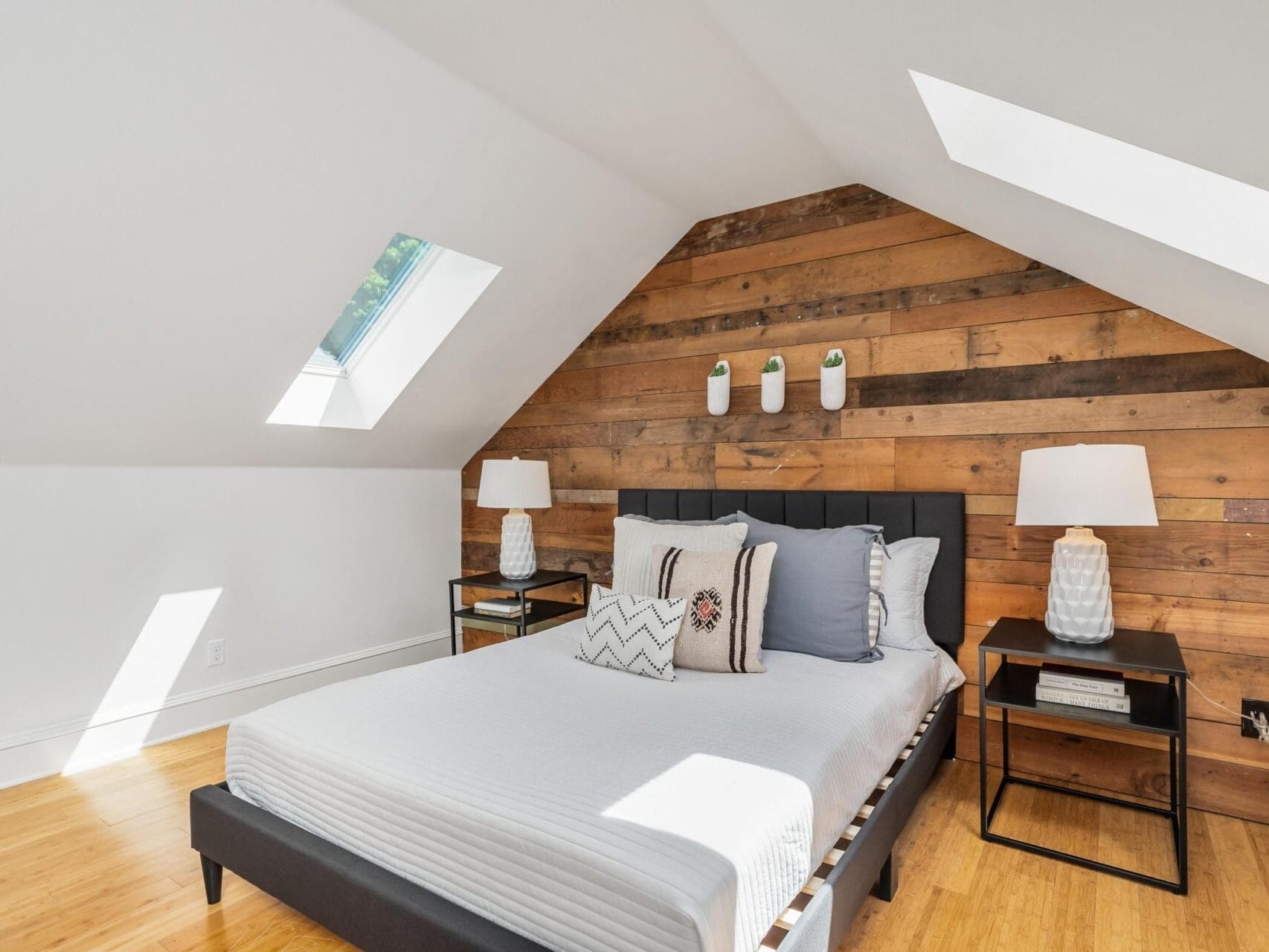 A cozy bedroom with a sloped ceiling features a bed with white and gray pillows, flanked by black side tables and white lamps. A rustic wooden accent wall enhances the space, and skylights provide natural light.