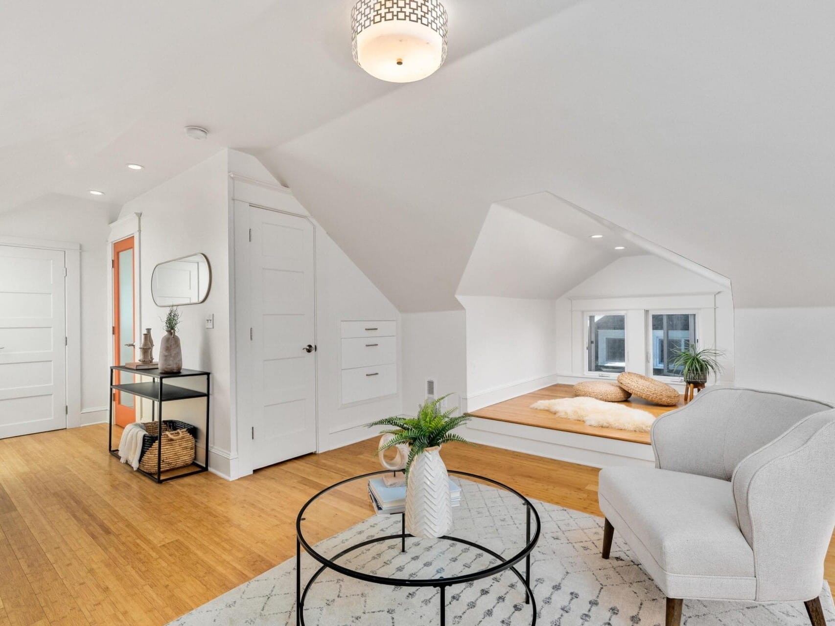 A bright, minimalist attic room with white walls and hardwood floors. A cozy seating area features a round glass table and upholstered chair. A built-in nook with cushions is by the window. A small console table with decor is against the wall.