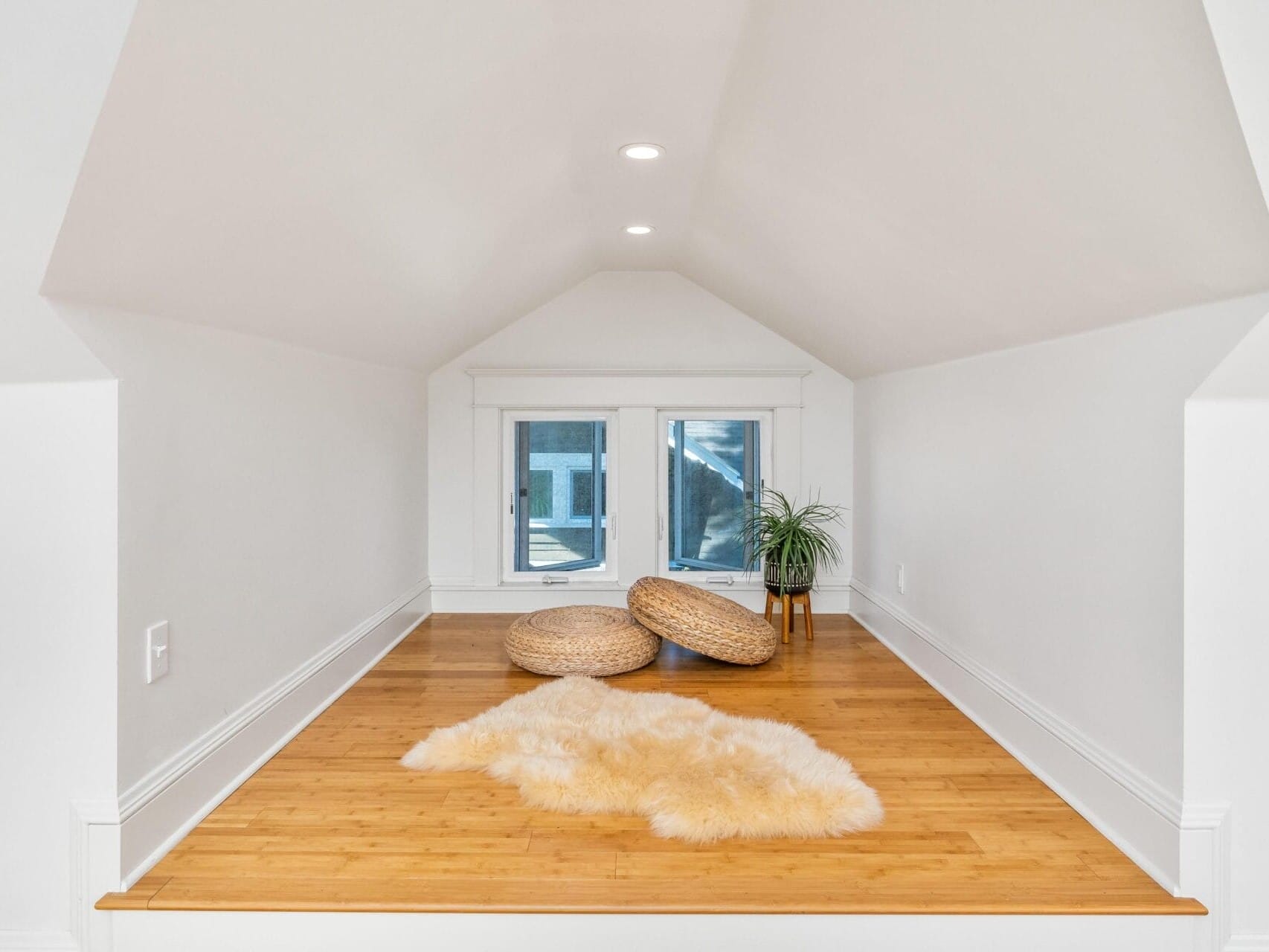 A cozy attic space with white walls and a wooden floor. Two round woven cushions and a fluffy cream rug are on the floor. A small plant is placed in the corner by a window, allowing natural light to enter.