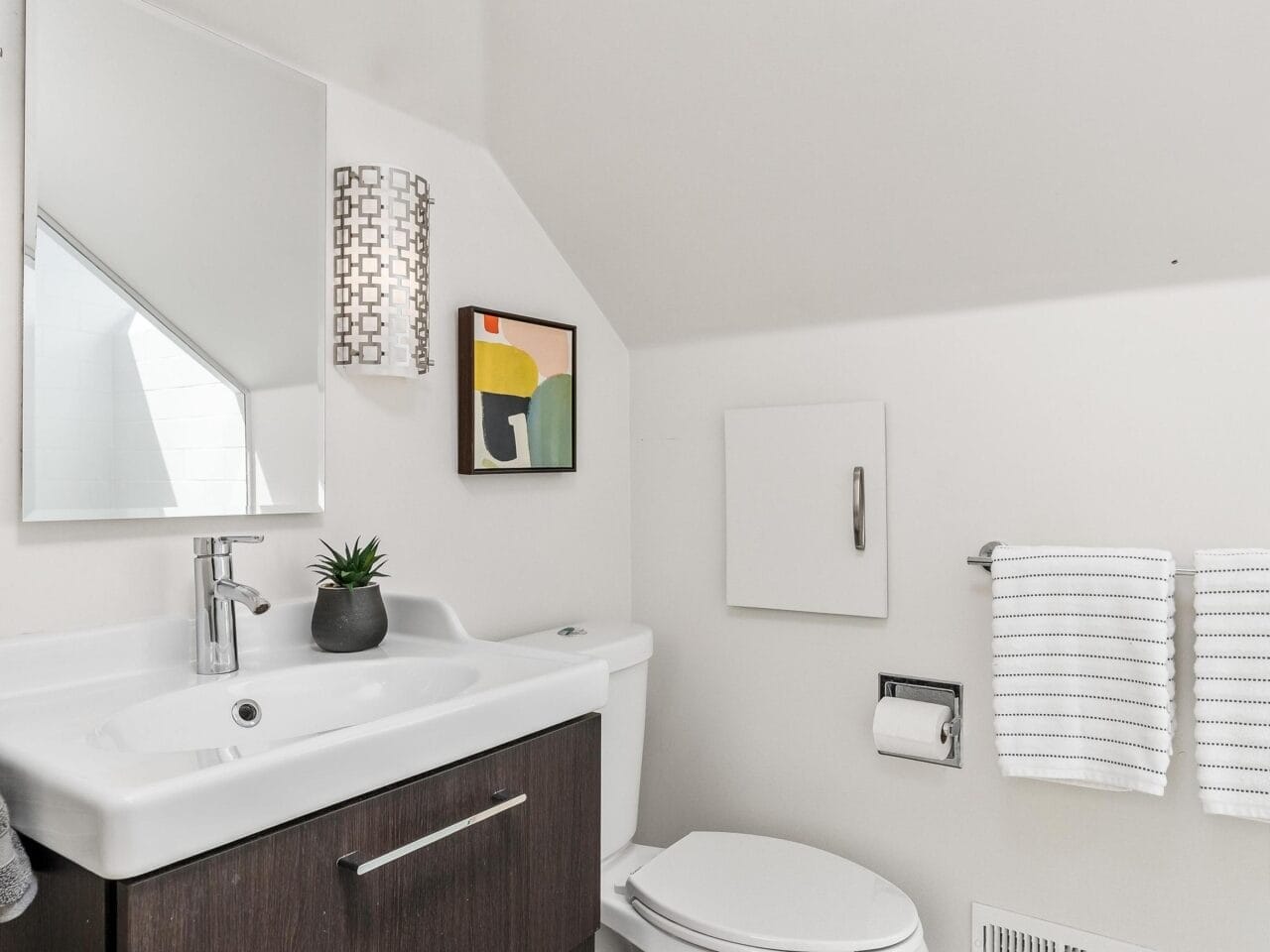 A modern bathroom featuring a white toilet, a dark wood vanity with a white sink, a small plant, and a wall-mounted mirror. White towels hang nearby, and a colorful abstract painting decorates the wall under sloped ceiling.