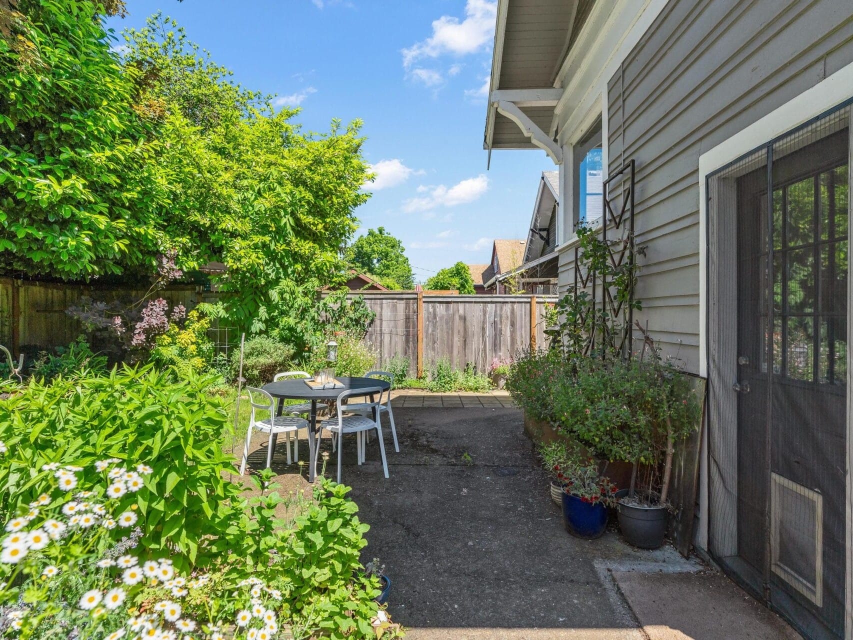 A cozy backyard patio with a wooden table and chairs surrounded by lush greenery, flowers, and potted plants. The patio is adjacent to a house with a screen door. A wooden fence encloses the area, under a clear blue sky.