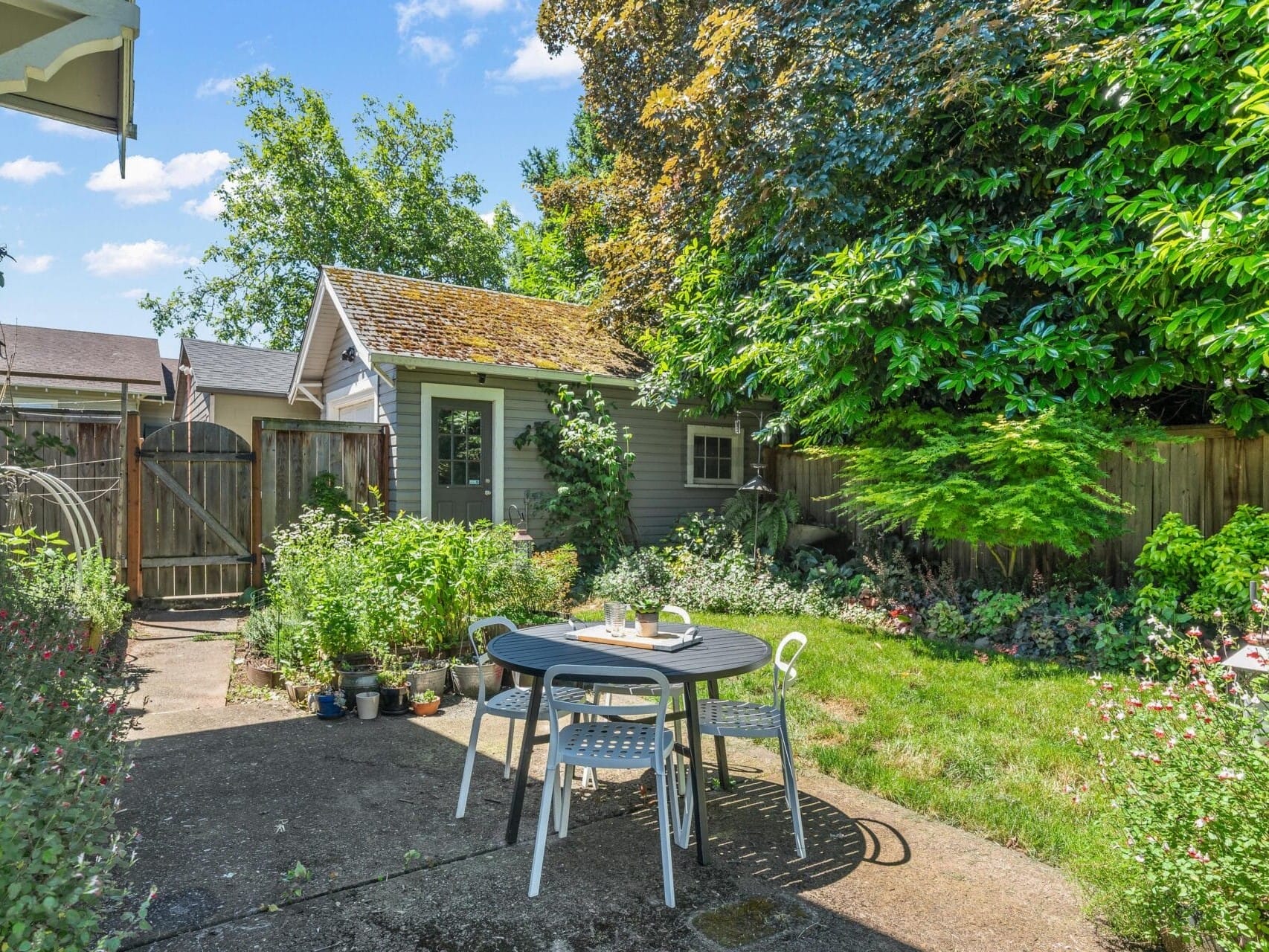 A cozy backyard garden with a round table and four chairs on a concrete patio. The area is surrounded by lush greenery, flowering plants, and a small garden shed. A wooden fence encloses the space on a sunny day.