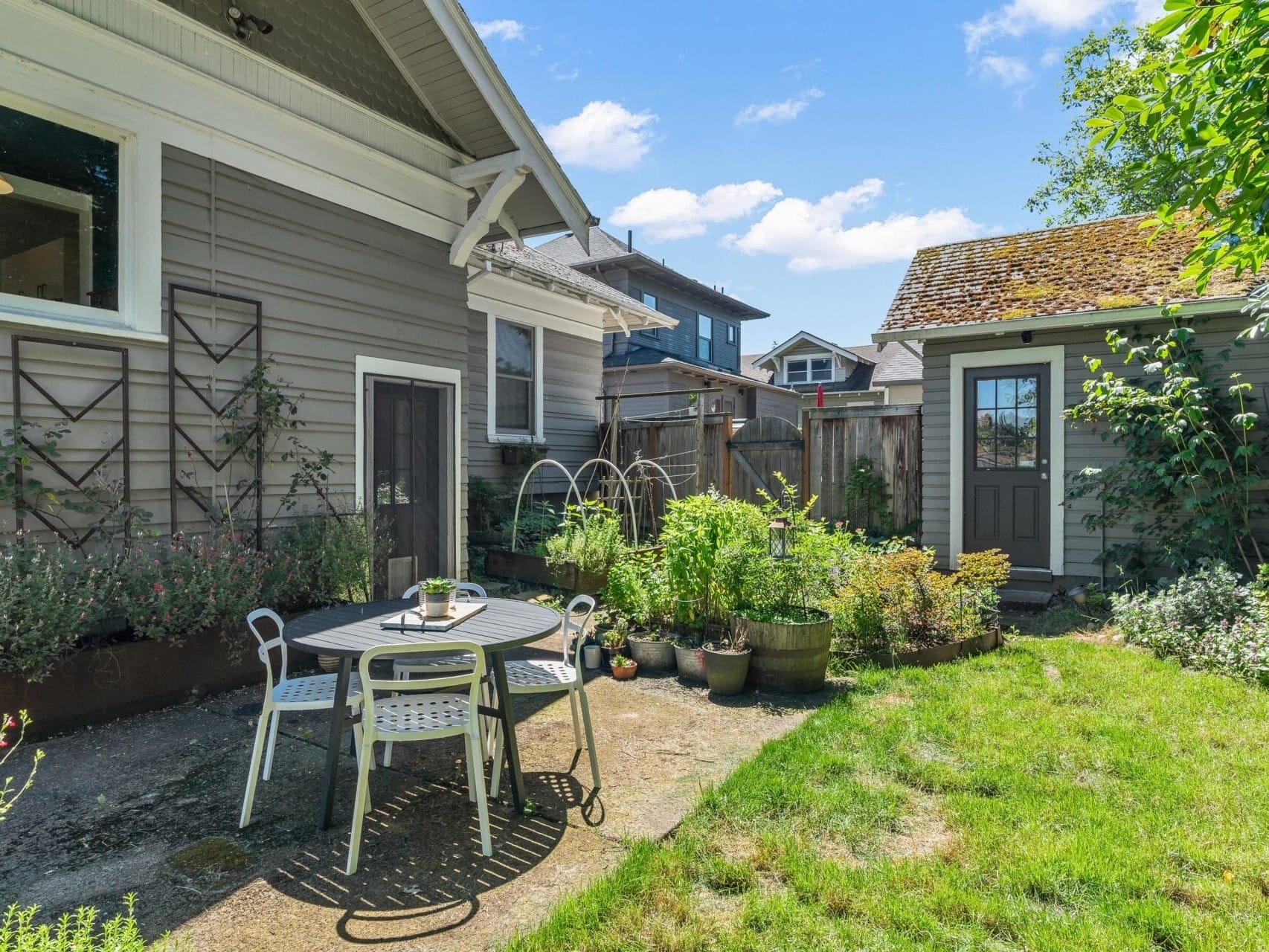 A small backyard garden with a patio table and chairs on a paved area. Surrounding the patio are various potted plants and garden beds. A wooden gate and additional small building are visible under a clear blue sky.