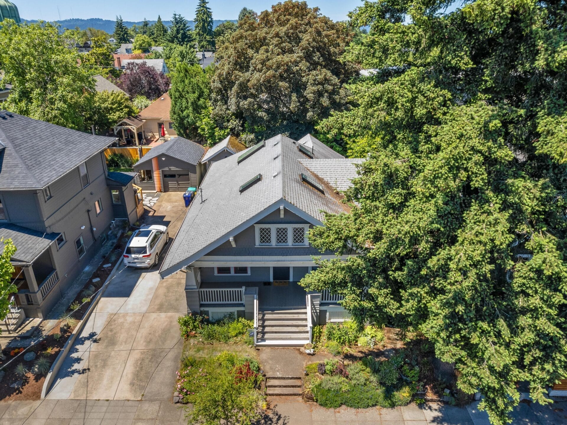 Aerial view of a gray, two-story house with a wide front porch and distinctive rooflines, surrounded by lush greenery. A driveway leads to a car parked beside the house. Neighboring houses are partially visible under a clear sky.