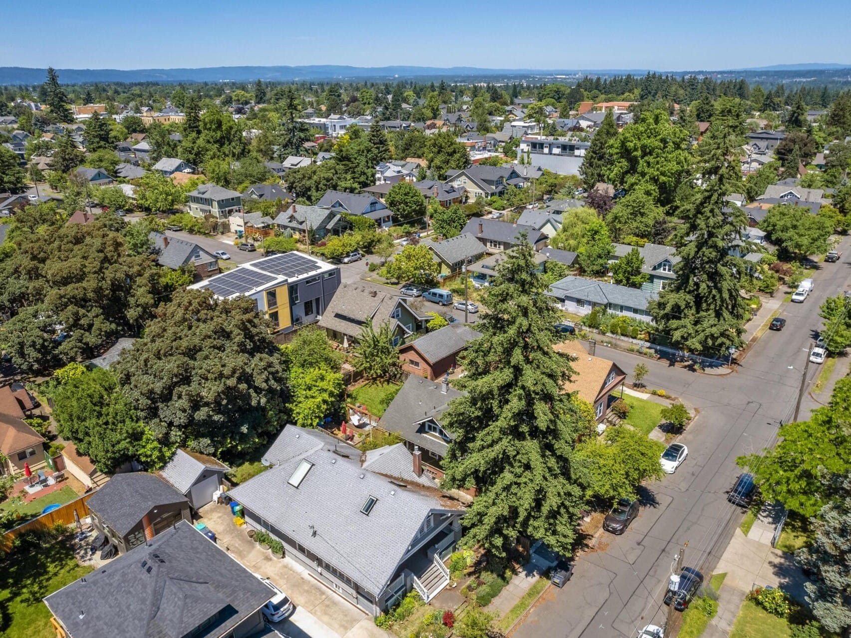 Aerial view of a suburban neighborhood with tree-lined streets and houses. Some homes have solar panels. The area is vibrant with greenery and a clear blue sky in the background. Cars are parked along the quiet residential streets.