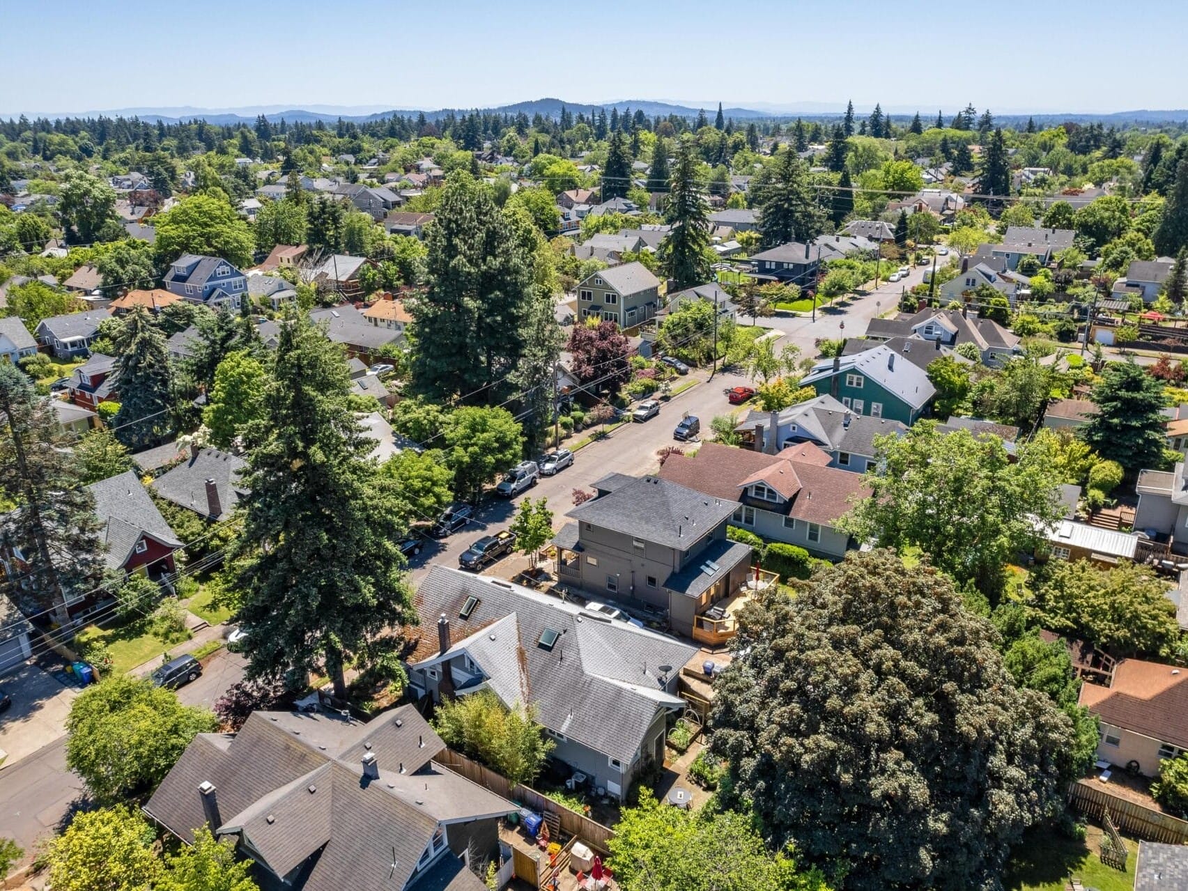 Aerial view of a suburban neighborhood with tree-lined streets and houses of various styles. The area is lush with greenery and surrounded by hills under a clear blue sky.