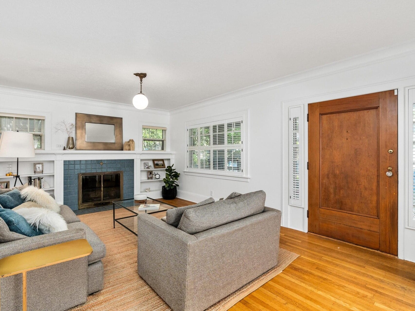 Cozy living room with two gray sofas, wooden coffee table, and a fireplace with a blue-tiled surround. A mirror and decor items are on the mantel. Large wooden door and windows with white shutters offer natural light. Warm-toned rug on hardwood floor.