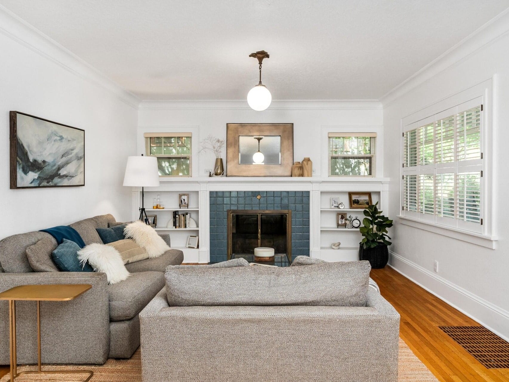 A cozy living room with gray sofas and a blue-tiled fireplace. A round light hangs above, and walls are decorated with art and a mirror. Shelves flank the fireplace, and a plant sits next to a window with white blinds.