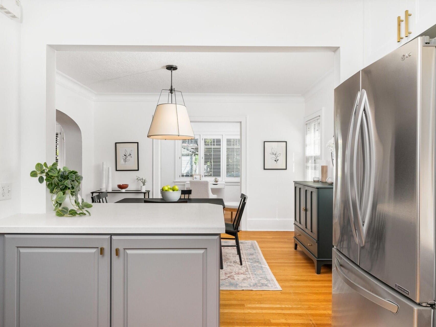 A modern kitchen with gray cabinets and a stainless steel refrigerator. A dining area with a table, black chairs, and a pendant light is visible in the background. The room features white walls, framed art, and a plant on the counter for decor.