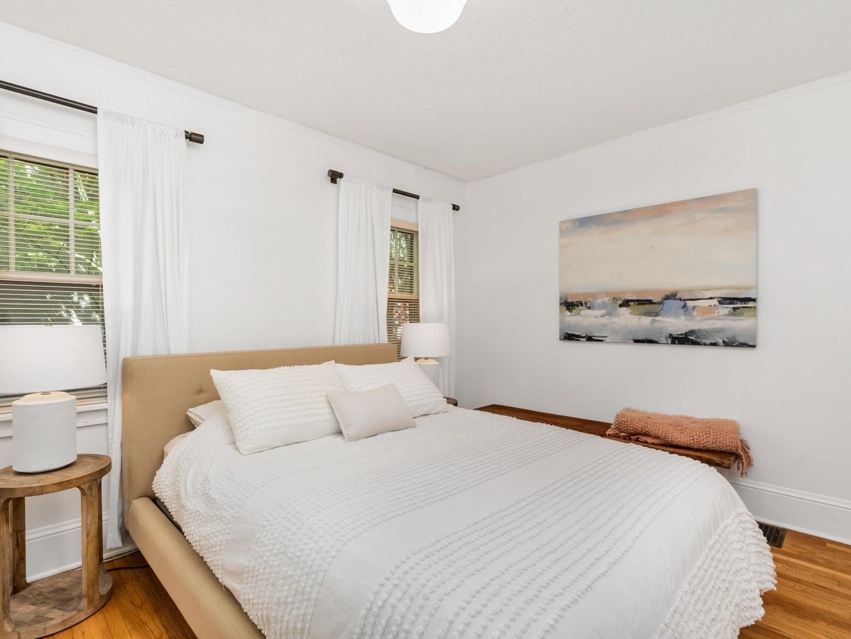 A cozy bedroom with a beige bed, white bedding, and a landscape painting on the wall. The room has two windows with white curtains, wooden floors, and a small wooden side table with a lamp. A soft blanket is draped over the beds edge.