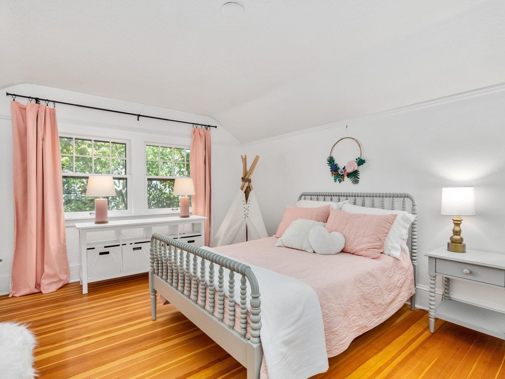 A cozy bedroom with a wooden floor, featuring a bed with pink bedding and heart-shaped pillows. The room includes a small nightstand with a lamp, a white desk by the window with pink curtains, and a decorative wreath with a teepee in the corner.