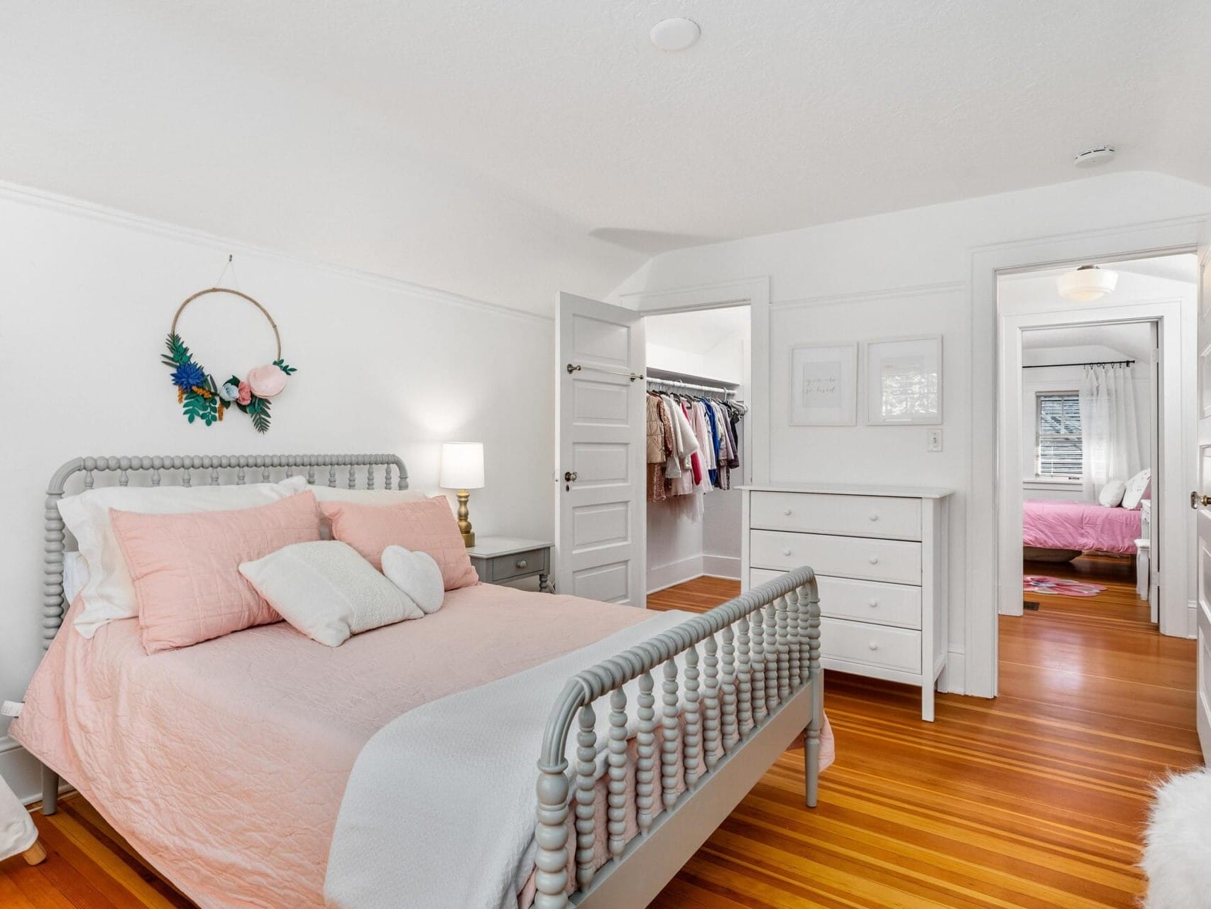 A bright bedroom with a white and light pink color scheme. It features a bed with pink bedding and pillows, a white dresser, and an open door revealing a closet. Wooden floors add warmth, and natural light streams in from a window in the next room.