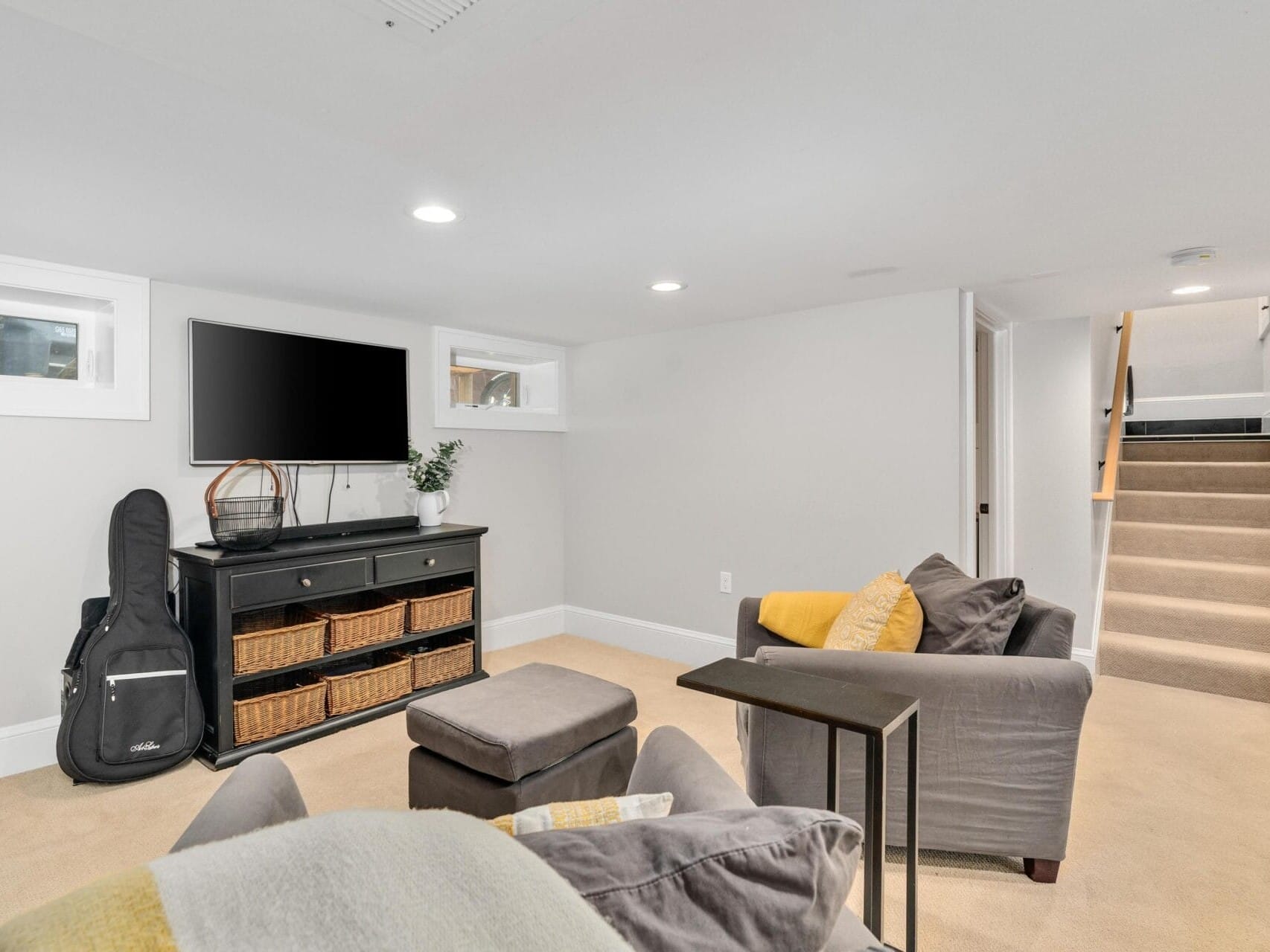 A cozy basement room with beige carpet, a black TV stand with a flat-screen TV, and storage baskets. Two gray armchairs with yellow and gray cushions face the TV. A guitar in a black case leans against the wall. Stairs lead up on the right.