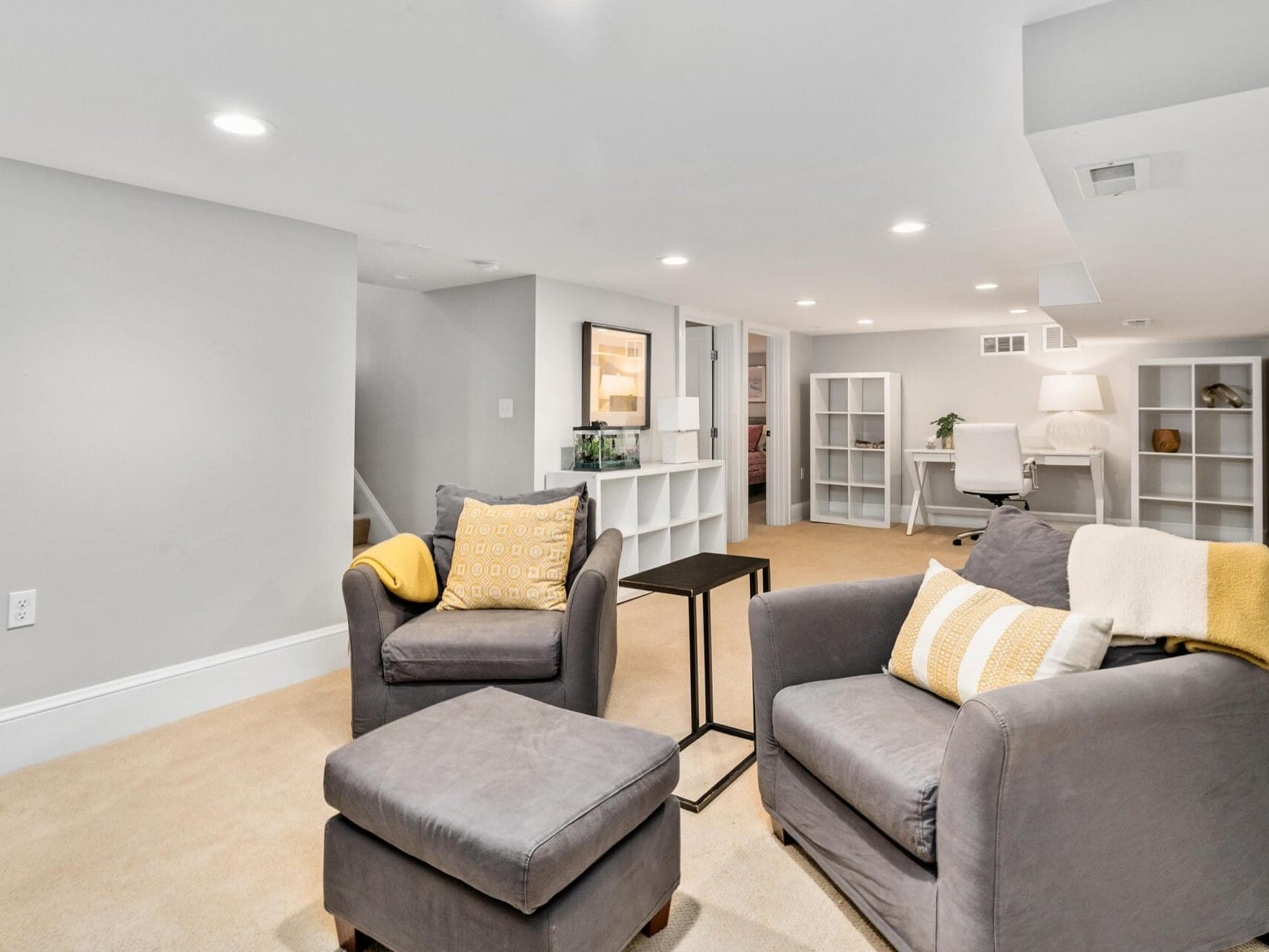 A cozy basement living space with two gray armchairs, one with a matching ottoman, decorated with yellow and white cushions. A small side table is between the chairs. The room features neutral-colored walls, carpet, and built-in shelves.