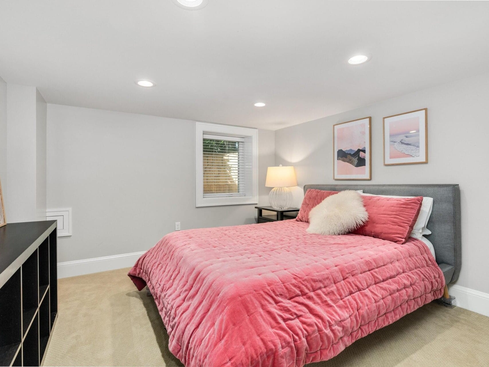 A cozy bedroom with a pink quilted bed, fluffy pillow, and a gray headboard. Theres a black storage unit on the left, a lamp on a nightstand, and three framed artworks on the walls. The room has beige carpet and recessed lighting.