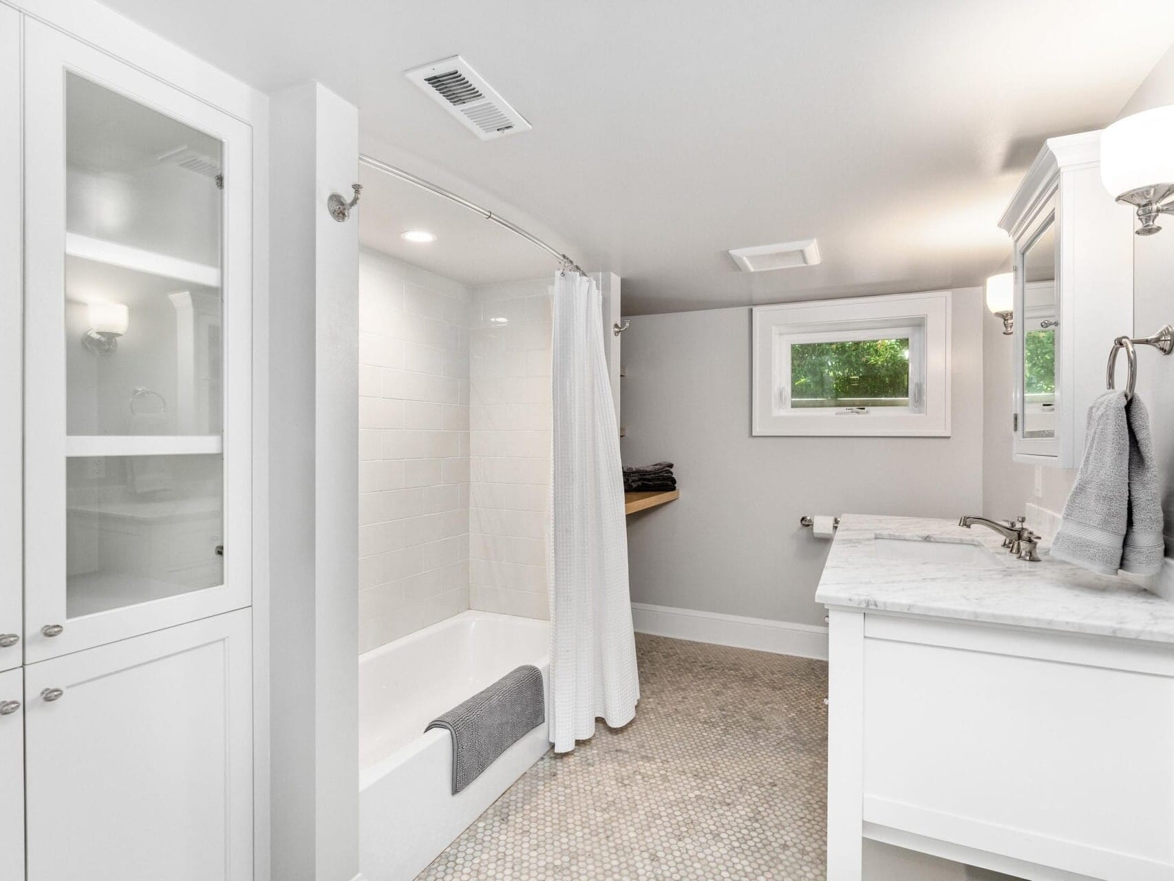 A bright, modern bathroom featuring a white bathtub with a shower curtain, a white vanity with a marble countertop and sink, and a glass cabinet. The room has light walls, a small window, and patterned flooring.