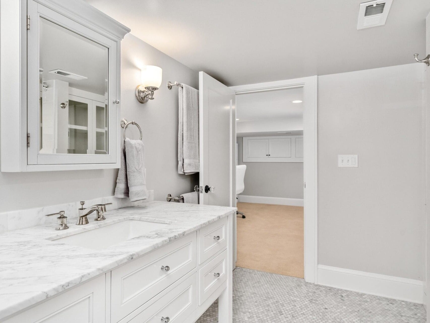 A modern bathroom with a marble countertop, double sink vanity, wall-mounted lighting, and a mirror. White cabinetry and a towel rack are present, and a partial view of an adjoining carpeted room with a chair and desk is visible through an open door.