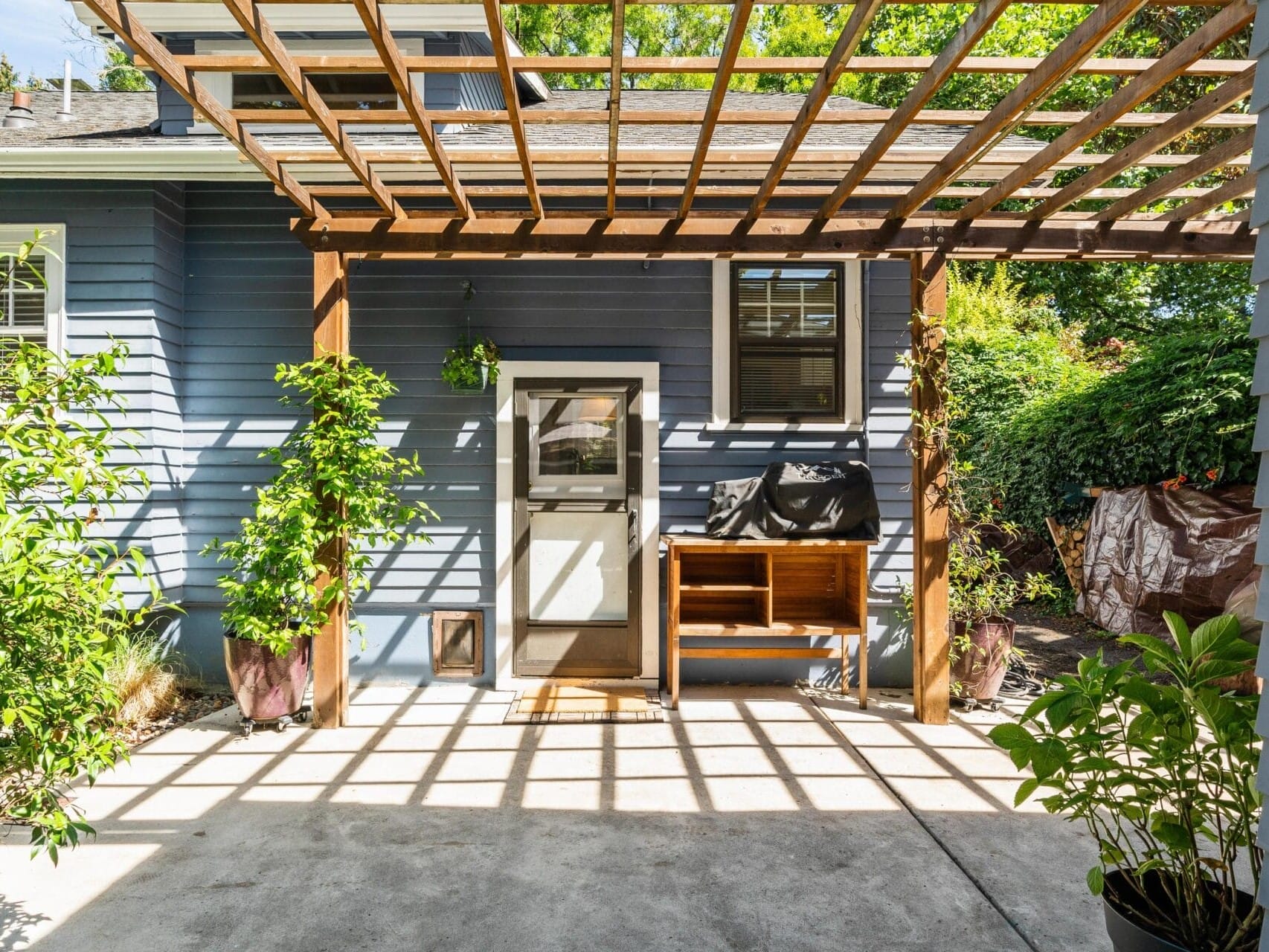 A sunny courtyard with a blue house featuring a white door. A wooden bench and grill sit under a wooden pergola casting shadows on the concrete floor. Potted plants line the edges, and leafy trees are visible in the background.