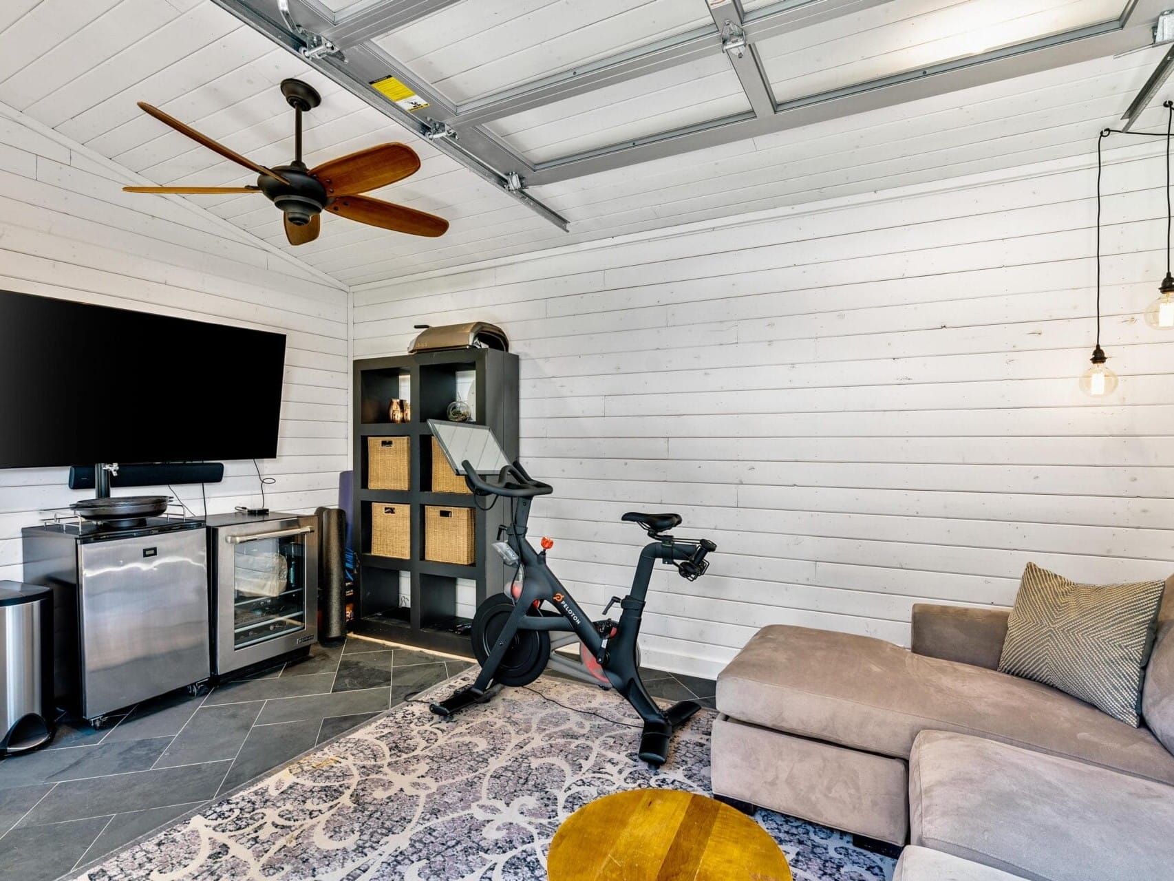 A home gym setup in a white paneled room with a stationary bike, a wall-mounted TV, and a compact shelving unit. Theres a beige sectional sofa, a rug, and two pendant lights hanging from the ceiling. A ceiling fan provides ventilation.