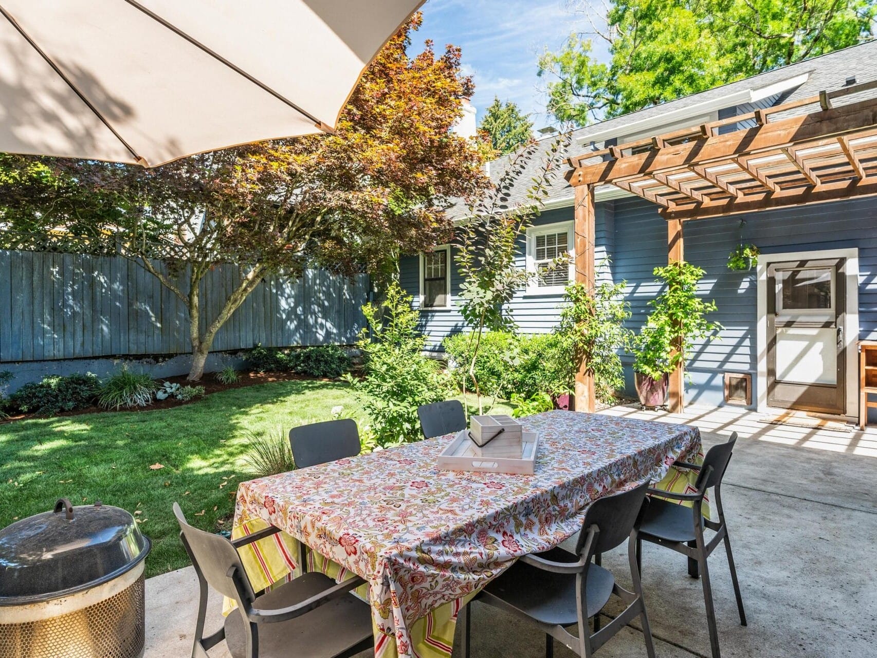 A backyard patio with a large table covered in a colorful tablecloth surrounded by chairs. A grill is on the left, and a pergola with climbing plants is attached to the house. Theres a tree and grass in the background under a blue sky.