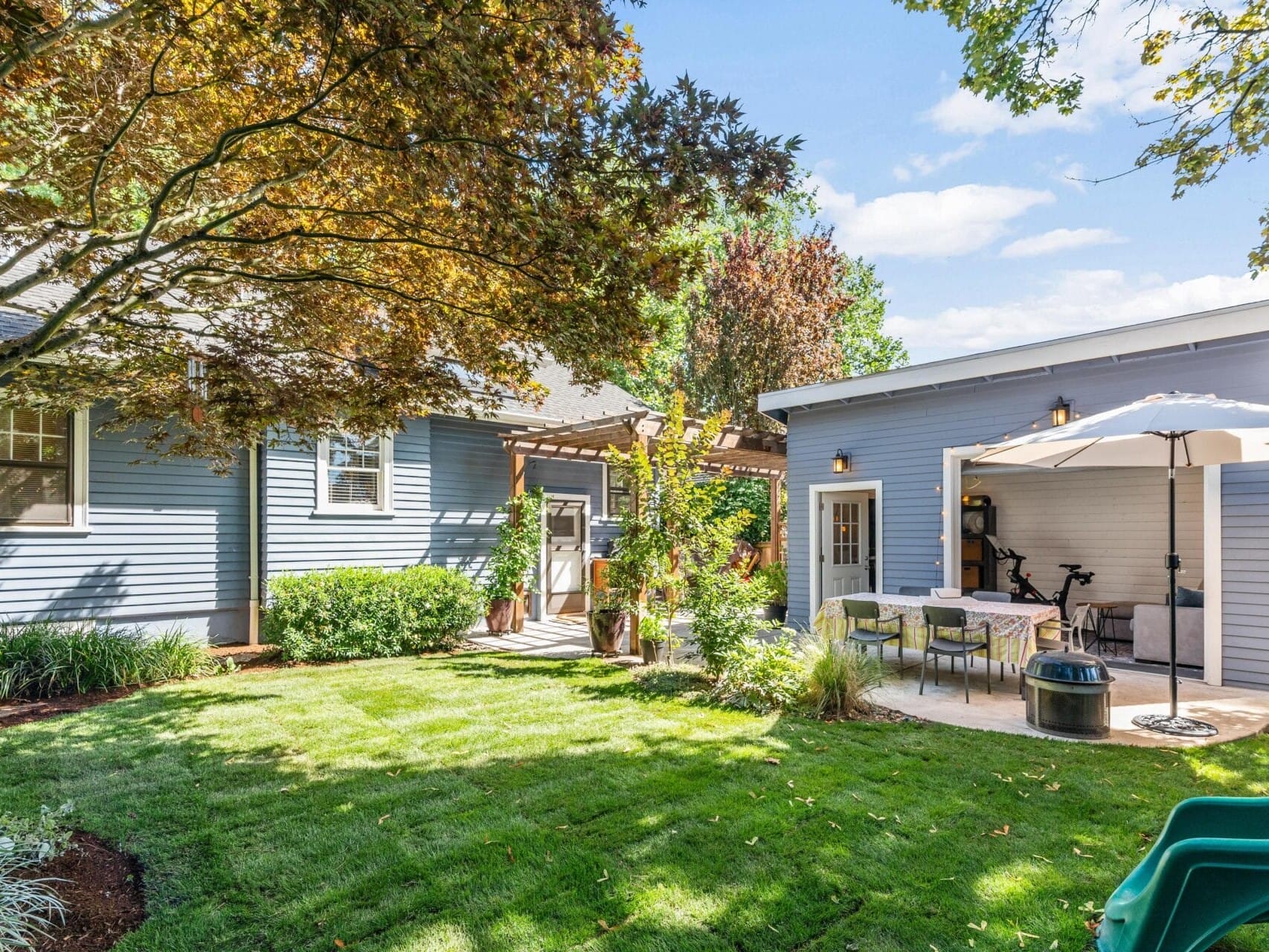 A sunny backyard with a green lawn, blue house, and attached patio. The patio has a table, chairs, and a large umbrella. Trees and plants provide shade and greenery. A small shed is in the background.