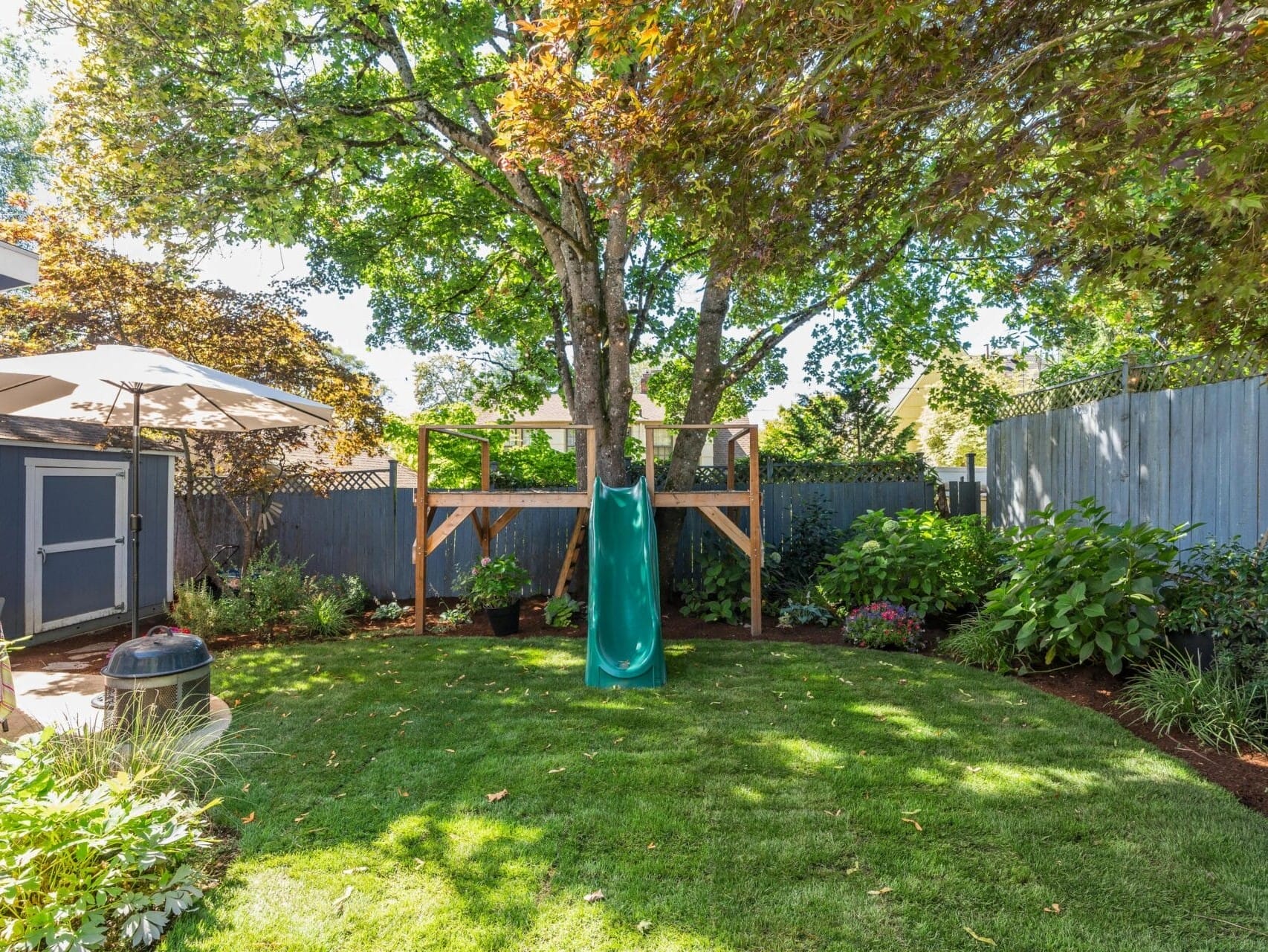 A backyard garden featuring a grassy lawn with a green slide attached to a wooden treehouse. Theres a patio umbrella on the left and various trees and plants surrounding the area, with a gray fence in the background.