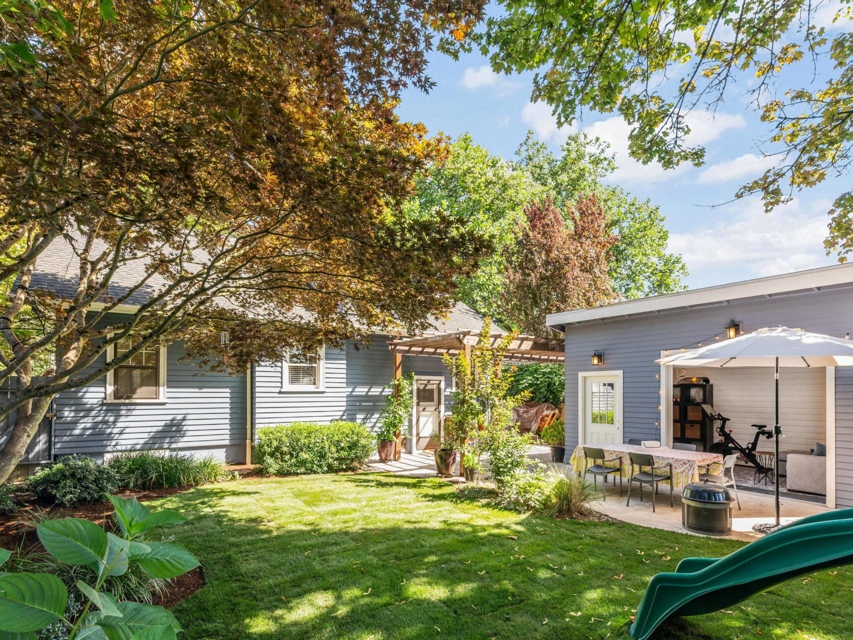 A lush backyard with a blue house and separate modern studio. A large tree provides shade over a grassy lawn. A patio with a table, chairs, and umbrella is nearby, and a green slide is visible on the right.