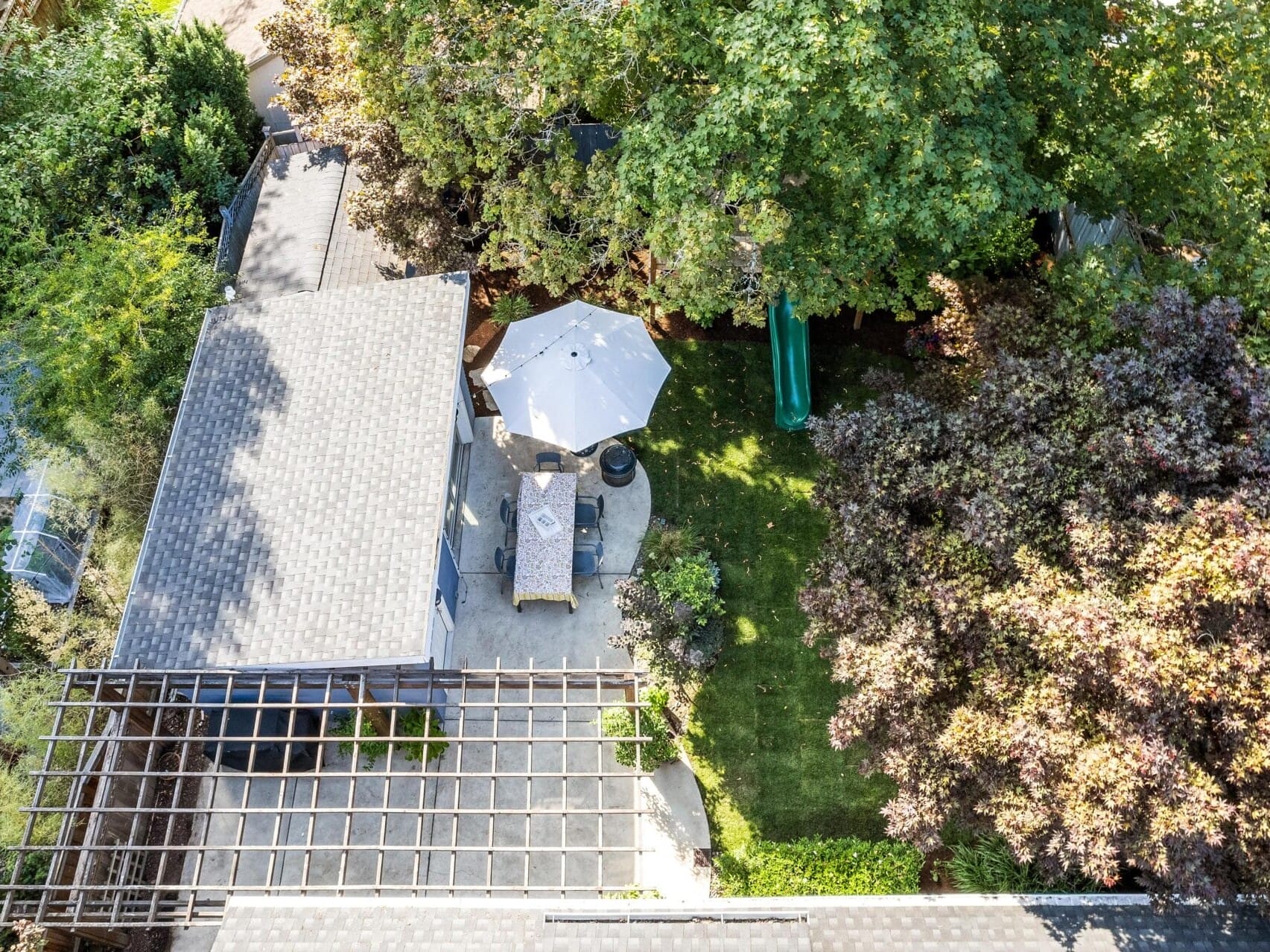 Aerial view of a backyard featuring a patio with a dining table and umbrella, a small grassy area, and surrounding trees. A pergola is attached to a gray-roofed structure on the left side.