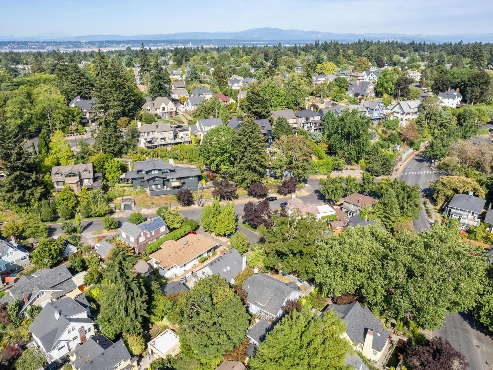 Aerial view of a suburban neighborhood with tree-lined streets and a variety of houses surrounded by greenery. Hills and distant mountains are visible under a clear blue sky.