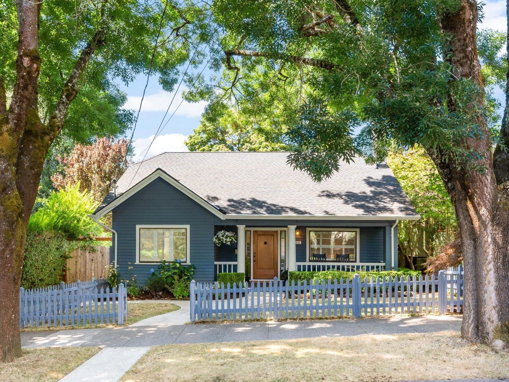 A charming blue cottage with a gray roof, surrounded by tall trees and a lush garden. It features a wooden front door, white-framed windows, and a blue picket fence with a concrete pathway leading to the entrance.