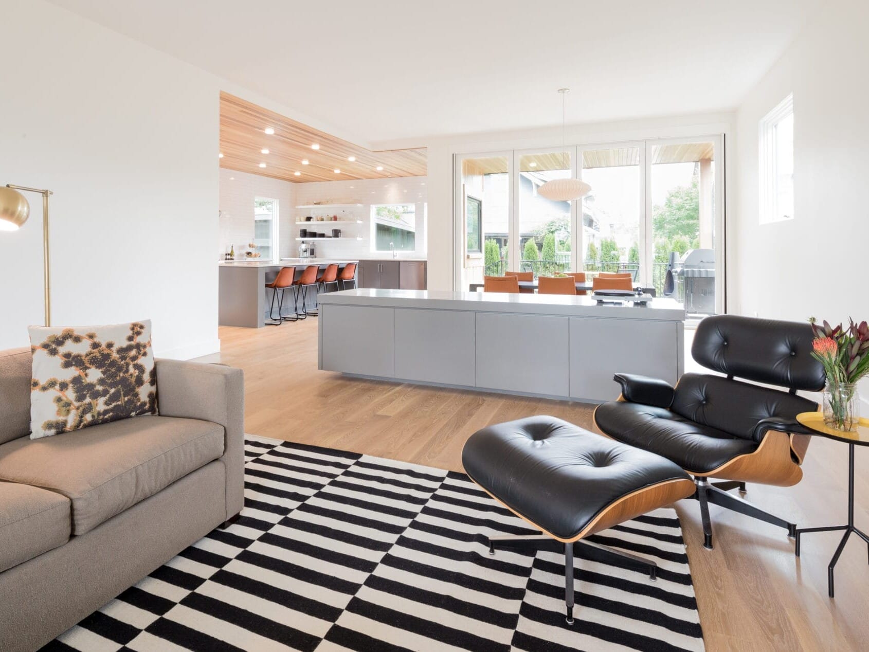 A modern living room in Portland, Oregon showcases a beige sofa with a patterned pillow, a black leather armchair and ottoman, and a striped rug. In the background, a minimalist kitchen with bar stools is visible, complemented by large windows that flood the space with natural light.