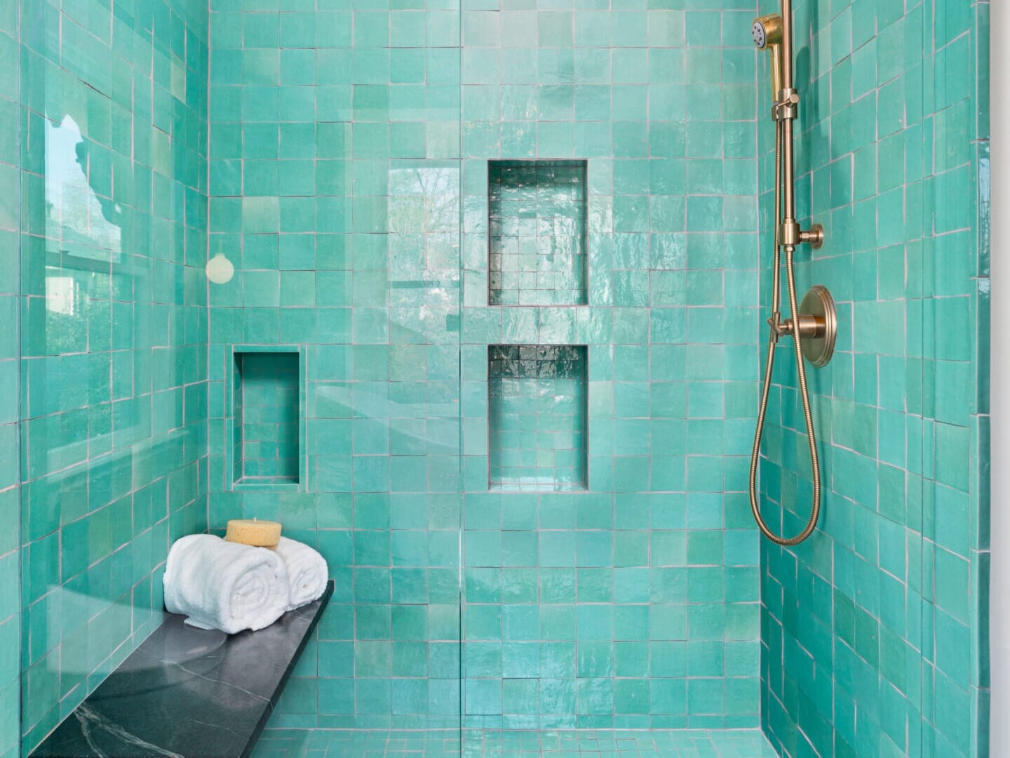 A modern shower in Portland, Oregon, showcasing glossy turquoise tiles and a gold rain showerhead. Two built-in niches complement the sleek design, while a black marble bench holds neatly folded white towels and a bar of soap.