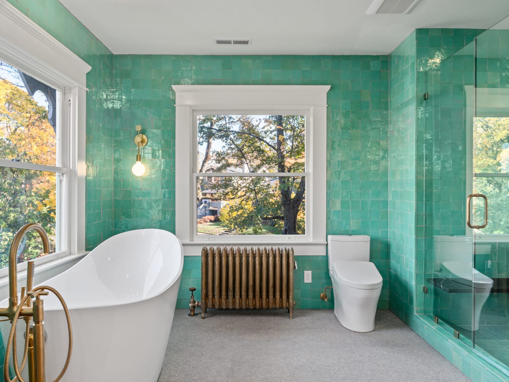 A bright bathroom in a Portland, Oregon home features teal tiled walls, a freestanding white bathtub, and a modern toilet. The glass shower enclosure and vintage-style radiator add charm. Large windows offer views of trees with autumn foliage, while a brass floor-mounted faucet accents the tub.