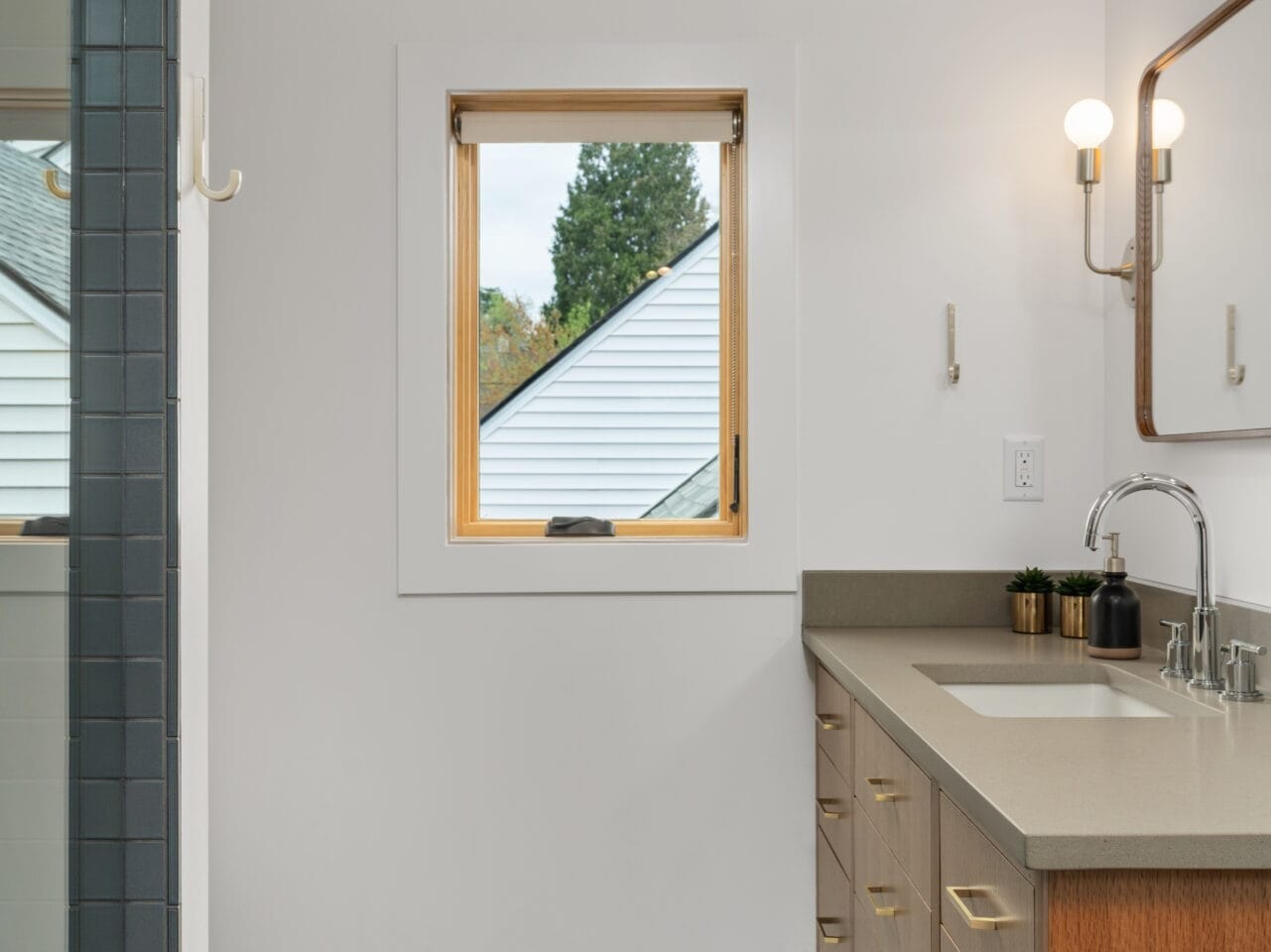 Modern bathroom in Portland, Oregon, featuring a wood-framed window, large mirror, and vanity with beige countertop and wooden drawers adorned with gold handles. A glass shower is partially visible on the left, while the floor showcases a dark, patterned design.