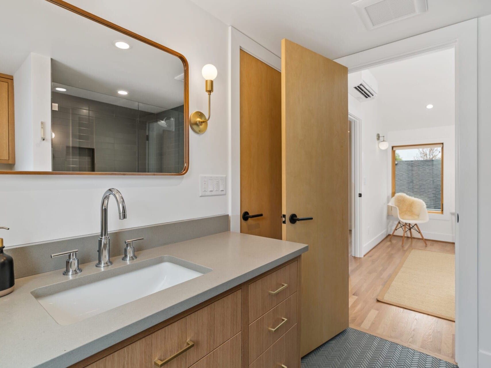 Modern bathroom with a light wooden vanity, gray countertop, and silver faucet. A large mirror and wall-mounted light fixture are above the sink. An open door leads to a bright Portland, Oregon room with a chair and rug. Wooden and glass accents are visible throughout.